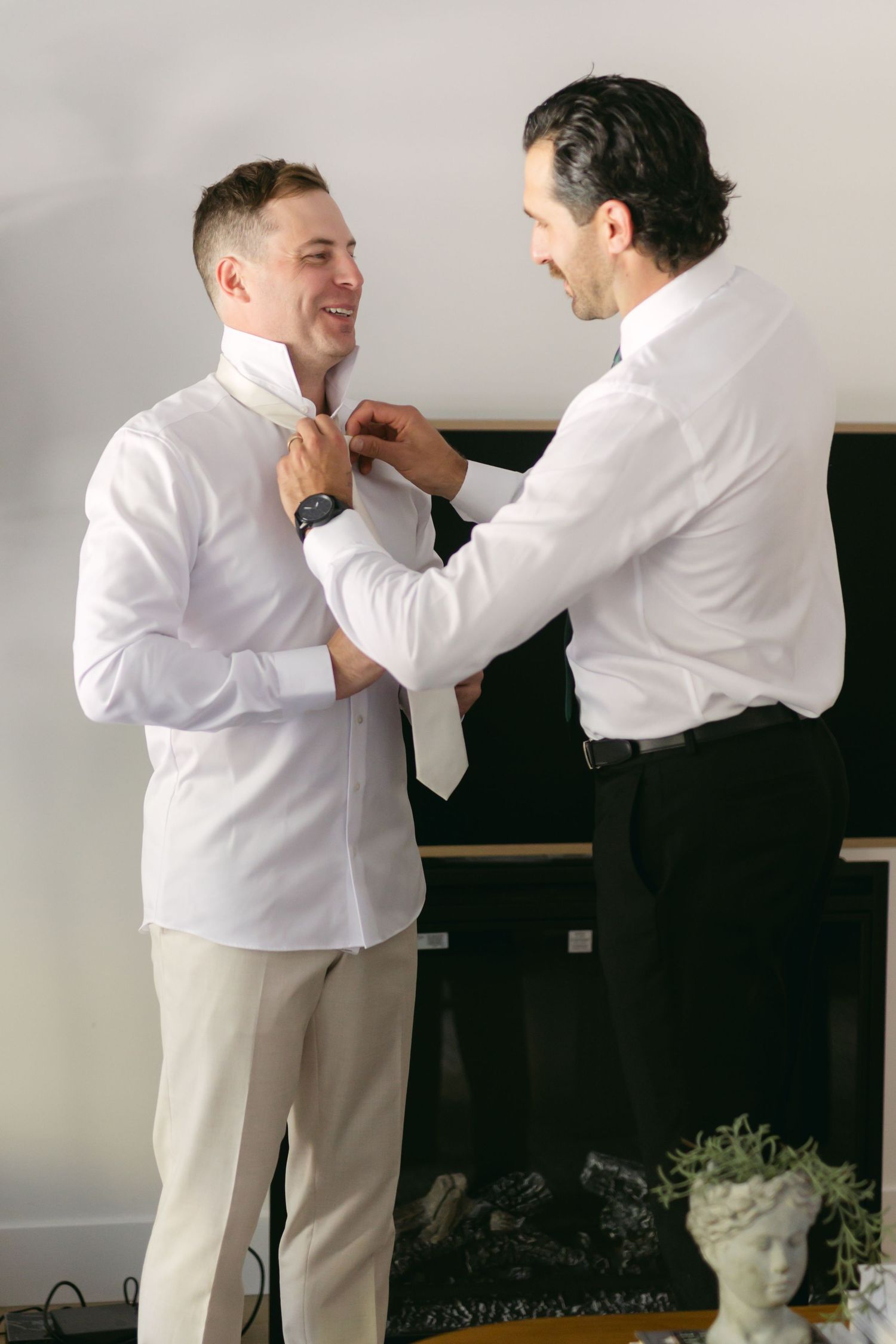 Two people in white shirts helping each other with formal attire preparations.
