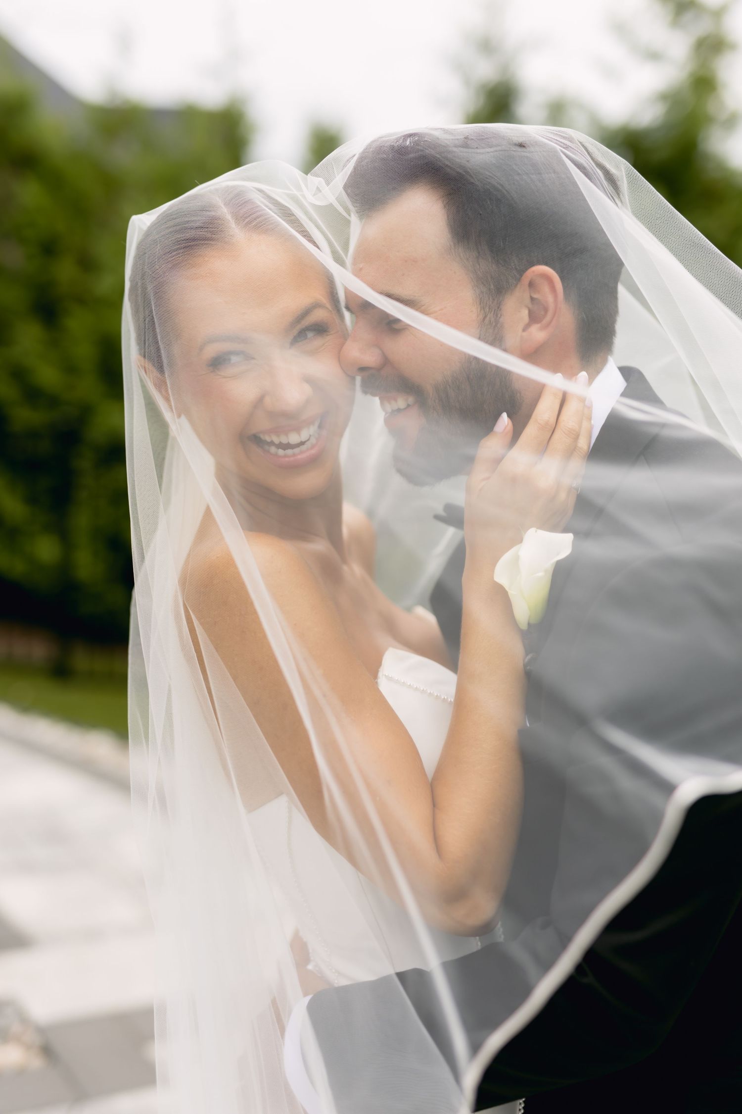 Laughing bride and groom share joyful moment together under flowing wedding veil outdoors.