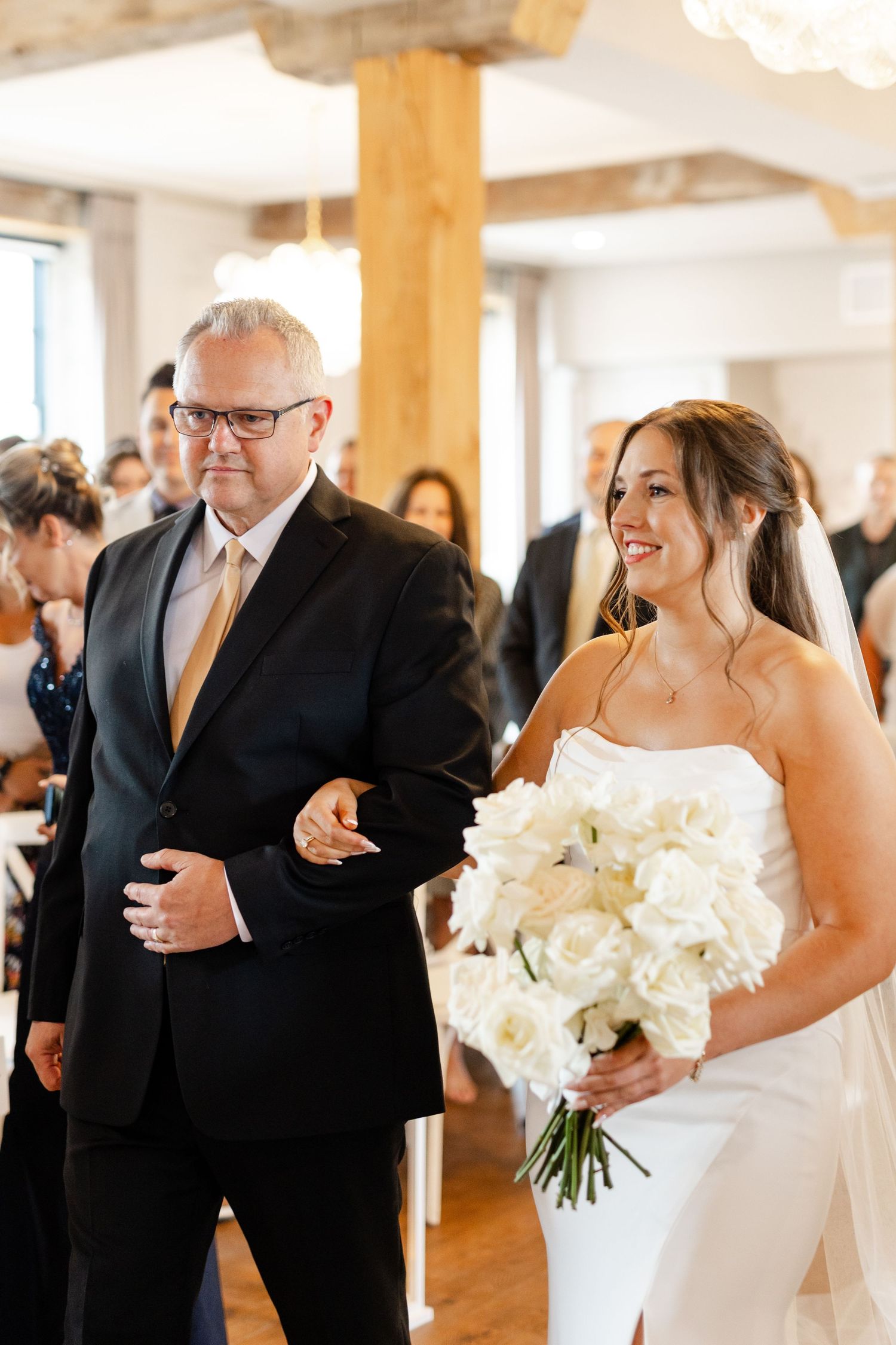 Bride walks down the aisle with her father holding a white rose bouquet in a rustic venue.