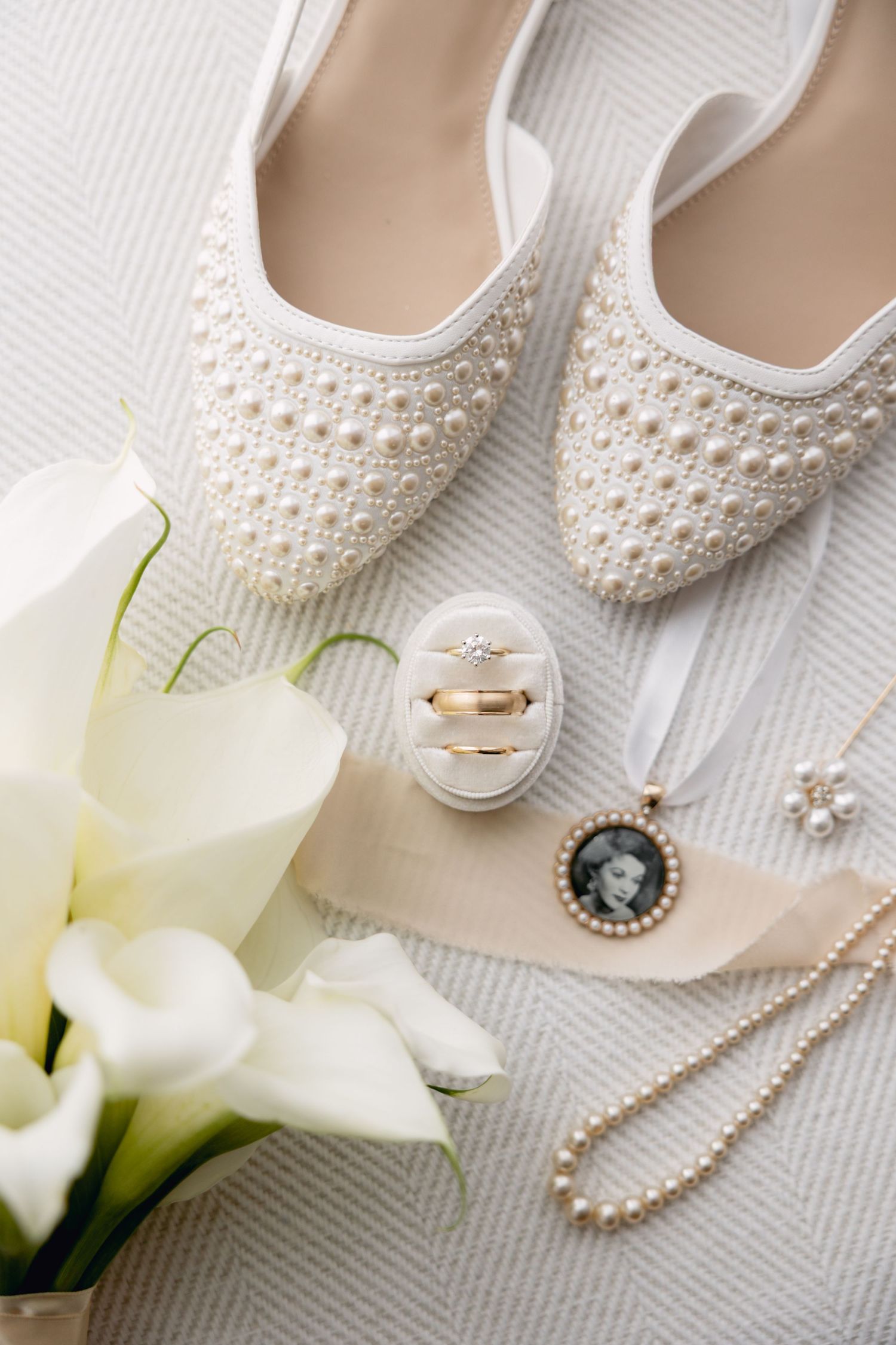 Bridal flat shoes adorned with pearls beside wedding rings, calla lilies, and pearl jewelry.