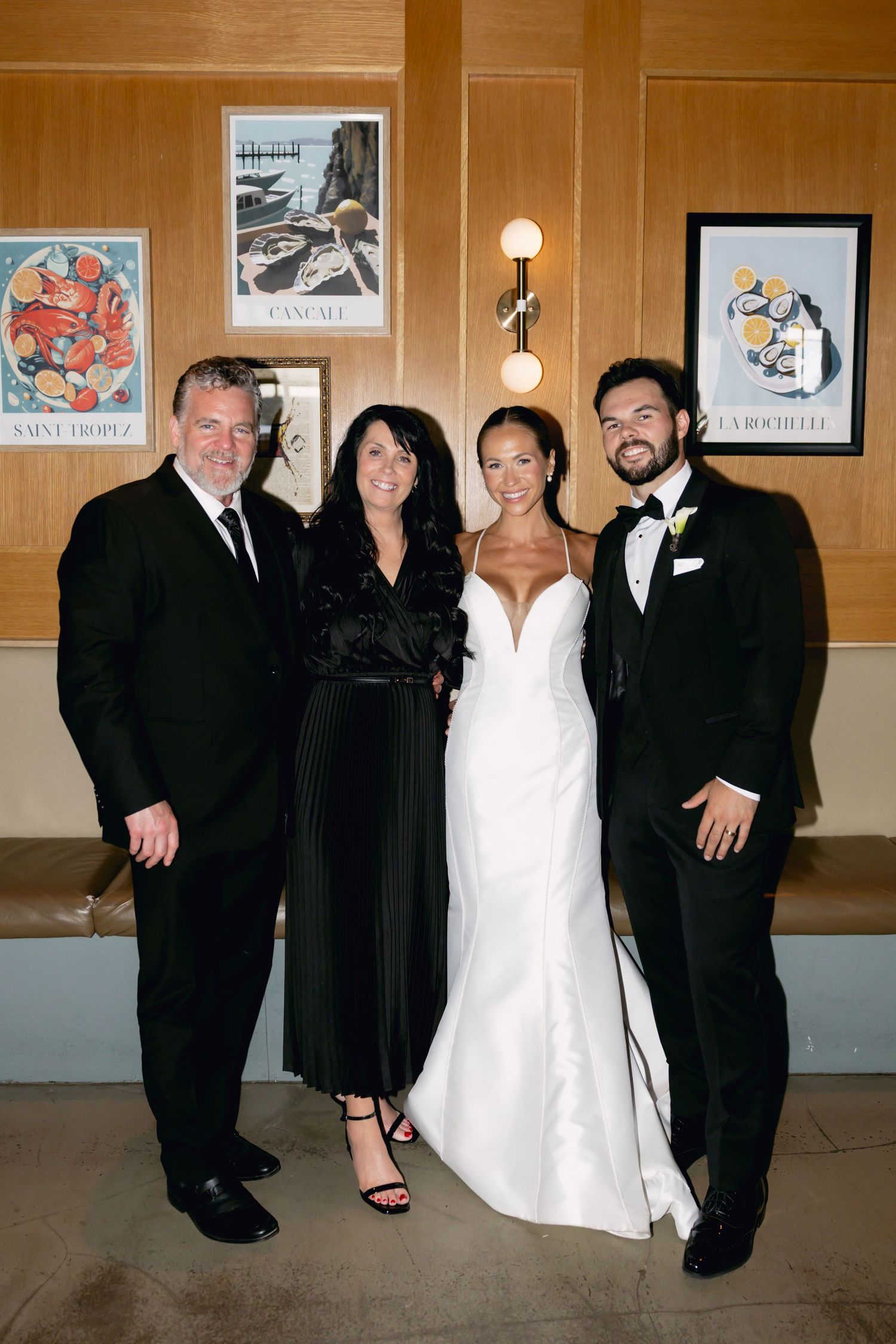 Bride and groom pose with two guests in front of French coastal art prints at reception.