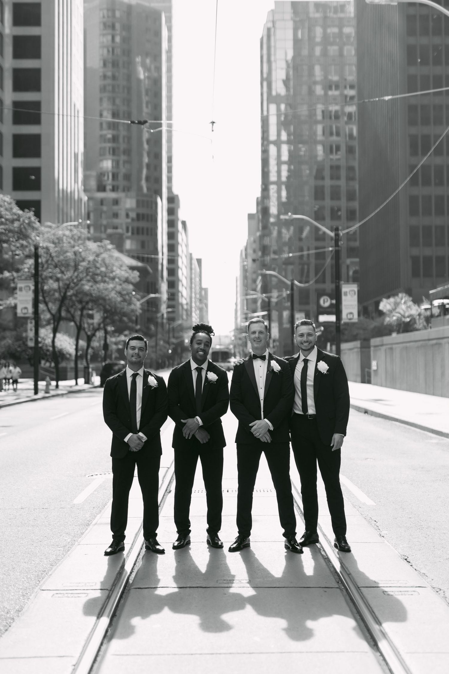 Groom and three groomsmen in black tuxedos posing on city street railway tracks in black and white photo.