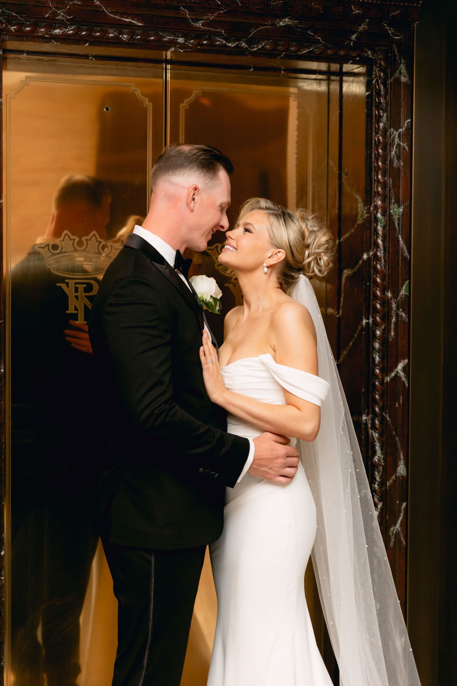 Bride and groom share romantic moment in front of ornate gold door with decorative monogram.
