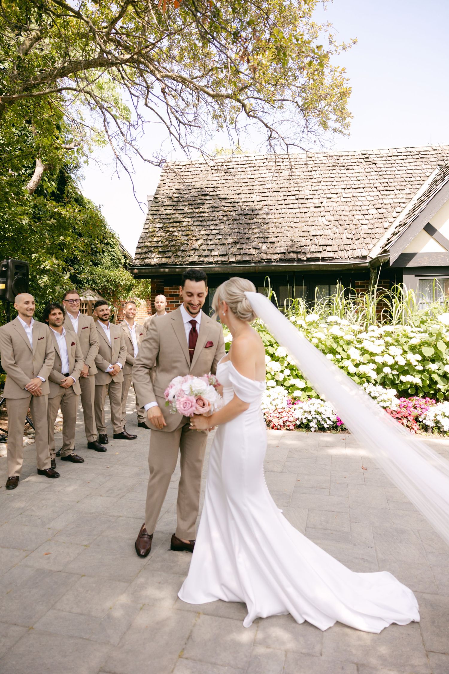 Wedding party in tan suits and white dress pose for formal photos in a garden setting.