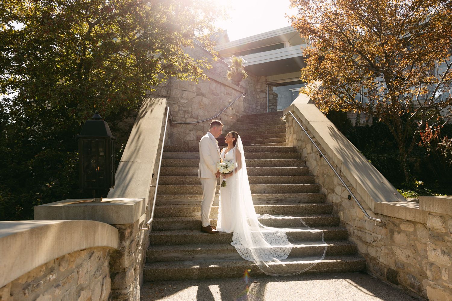 A couple in wedding attire poses on elegant stone steps during an outdoor autumn photoshoot.