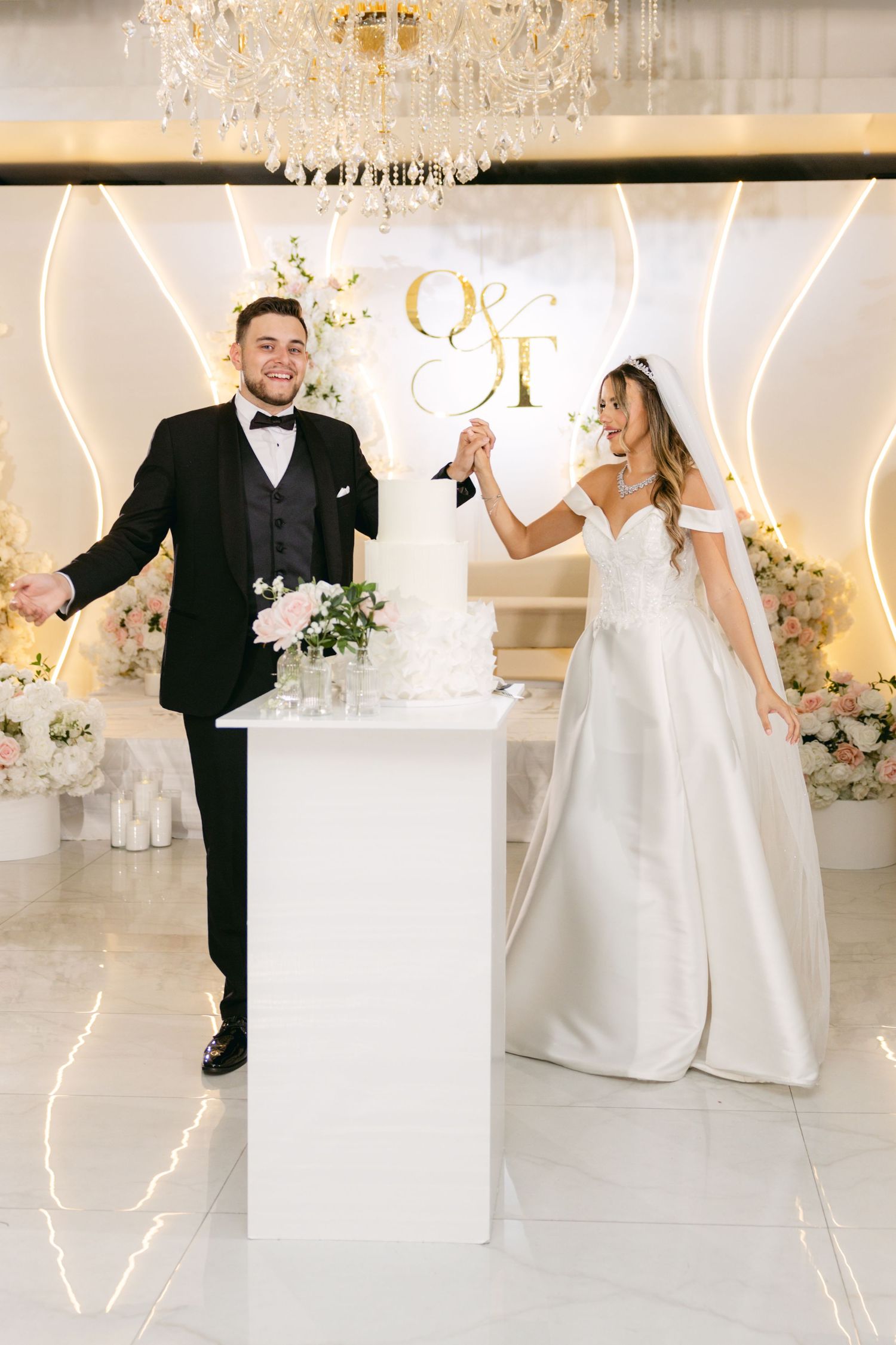 Newlyweds pose playfully by their wedding cake display with elegant white and gold decor.
