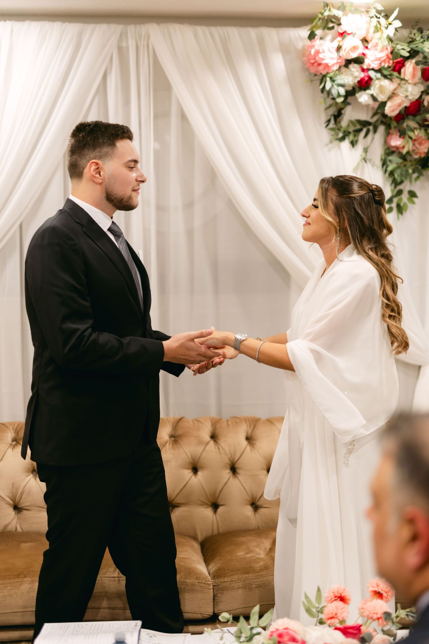 A couple holds hands during their wedding ceremony under white drapes and floral arrangements.