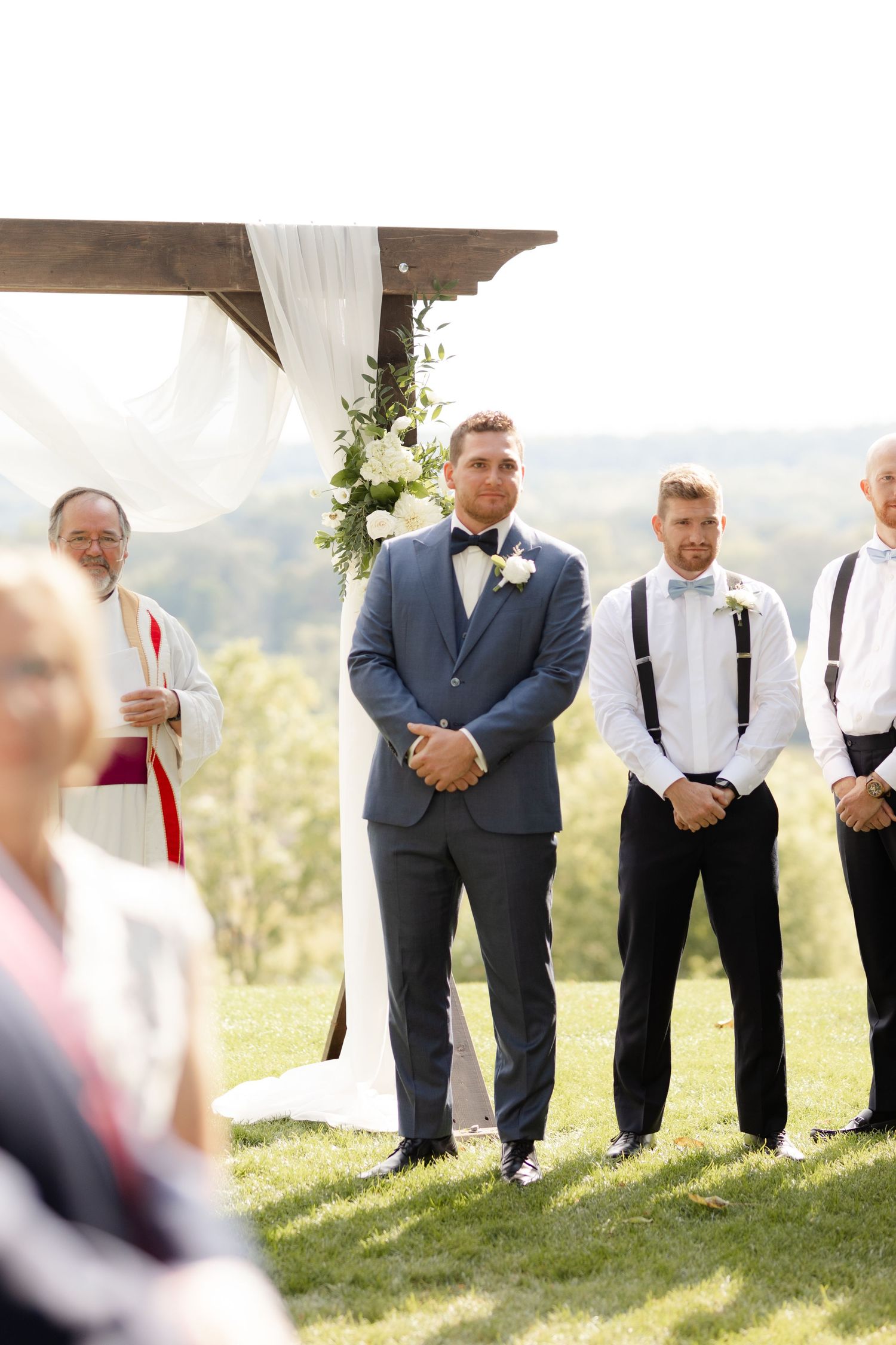 Wedding party members stand in line on grassy hill during outdoor ceremony.
