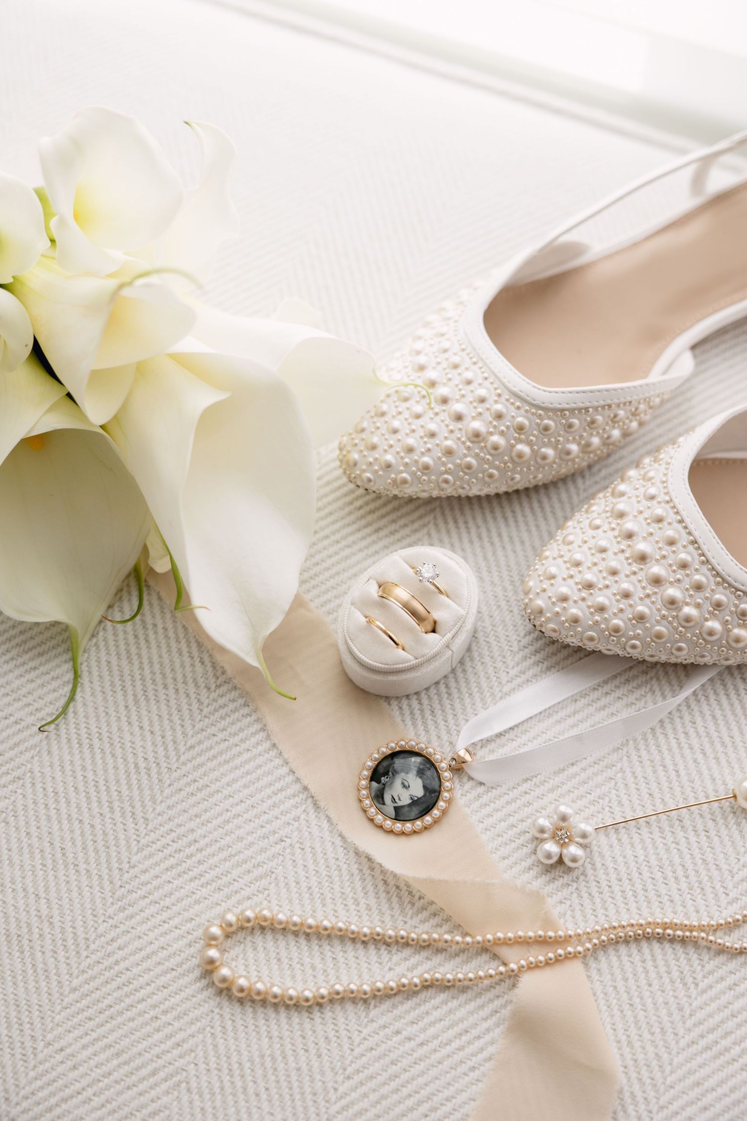 Pearl-embellished bridal flats, white calla lily bouquet, gold rings, pearl necklace, and locket arranged on white surface.