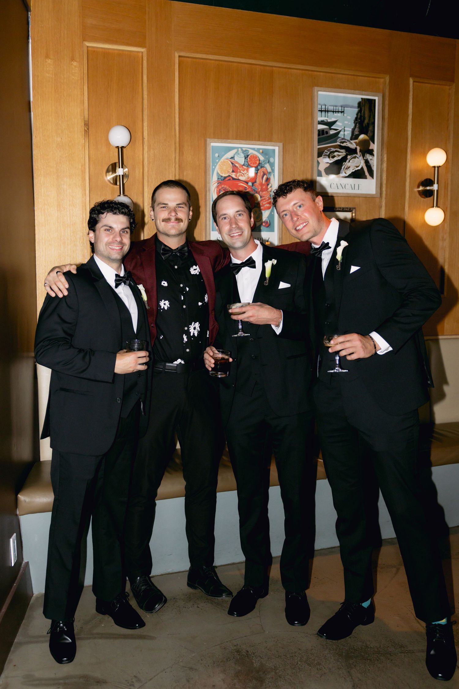 Four men in formal attire posing together holding drinks at a wedding reception.