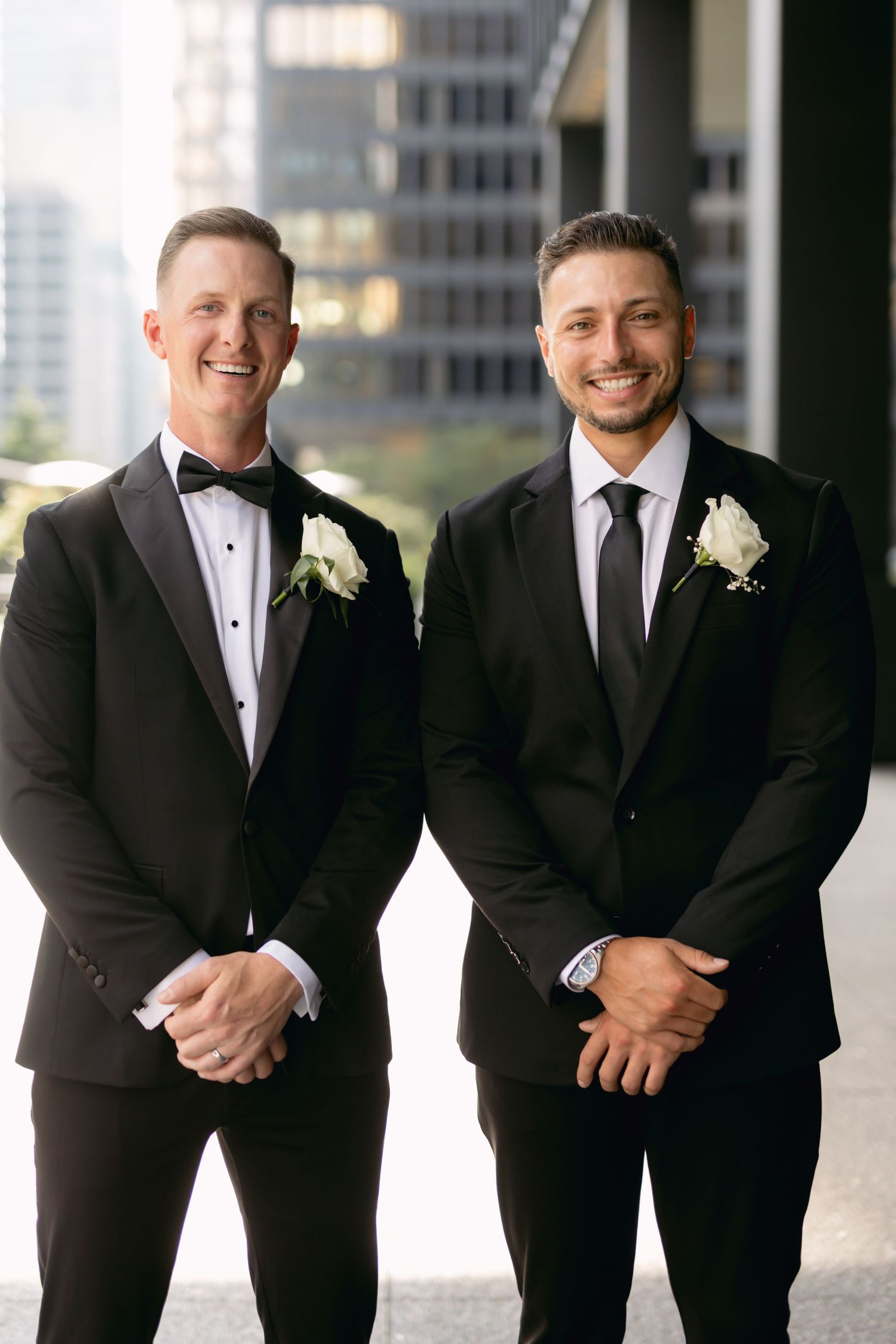 Two groomsmen in black suits with white boutonnieres smile outdoors with city buildings in background.