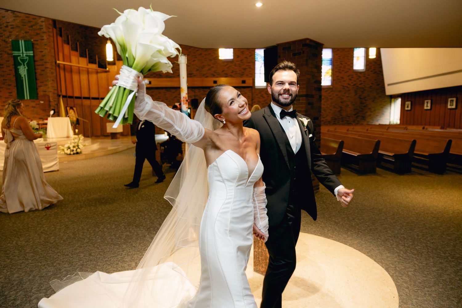 Joyful newlyweds celebrating recessional in church, bride raising bouquet with groom in tuxedo.