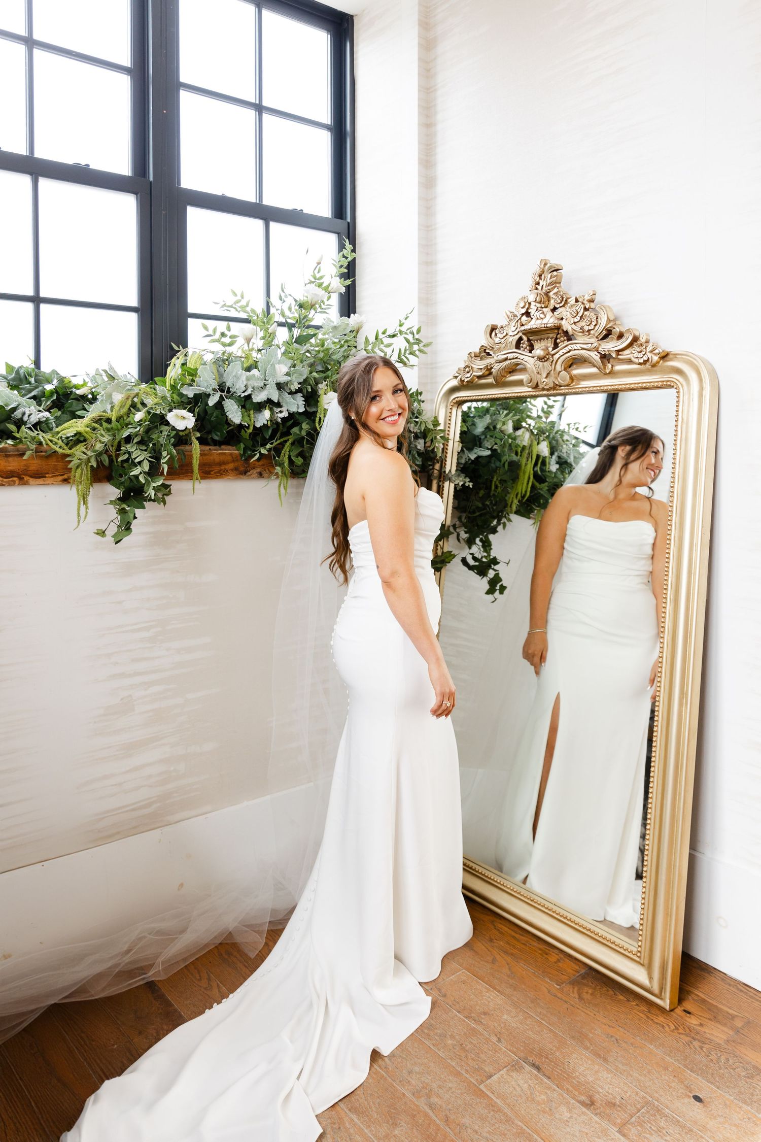 Smiling bride in sleek white gown poses beside an ornate gold mirror with floral greenery decor.