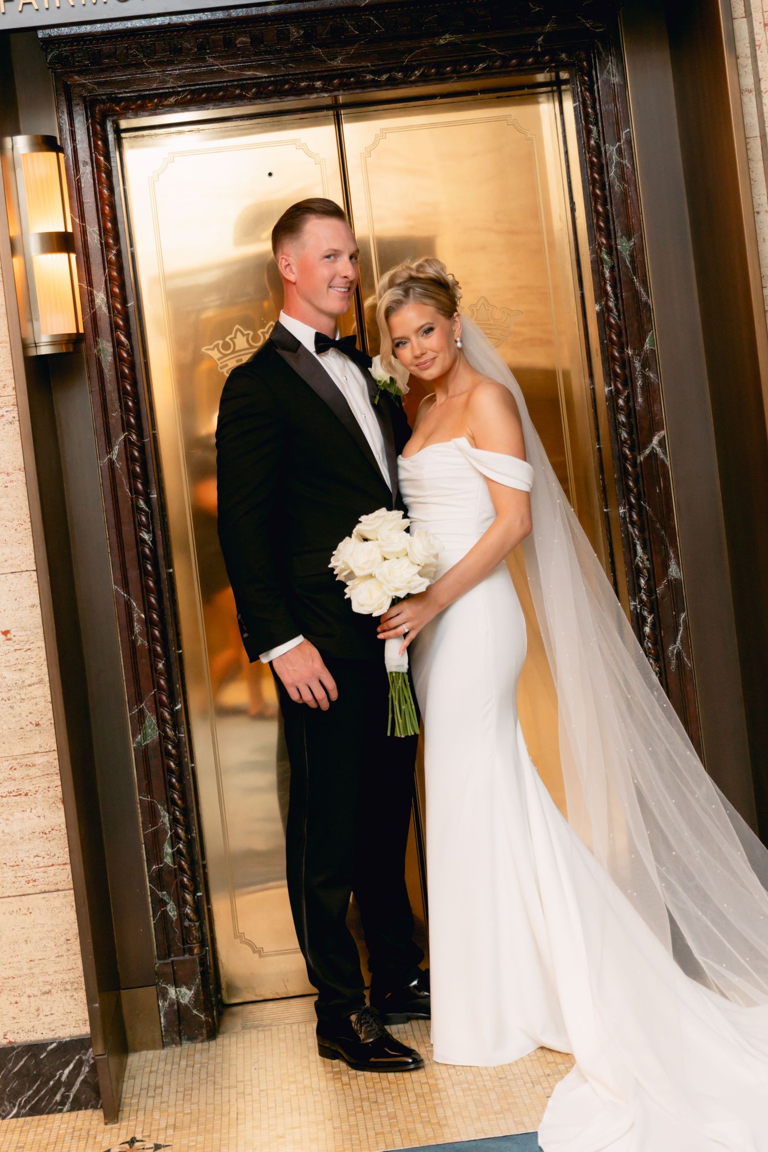 Elegant bride and groom pose in front of ornate gold elevator doors, bride holding white bouquet in fitted gown with veil.
