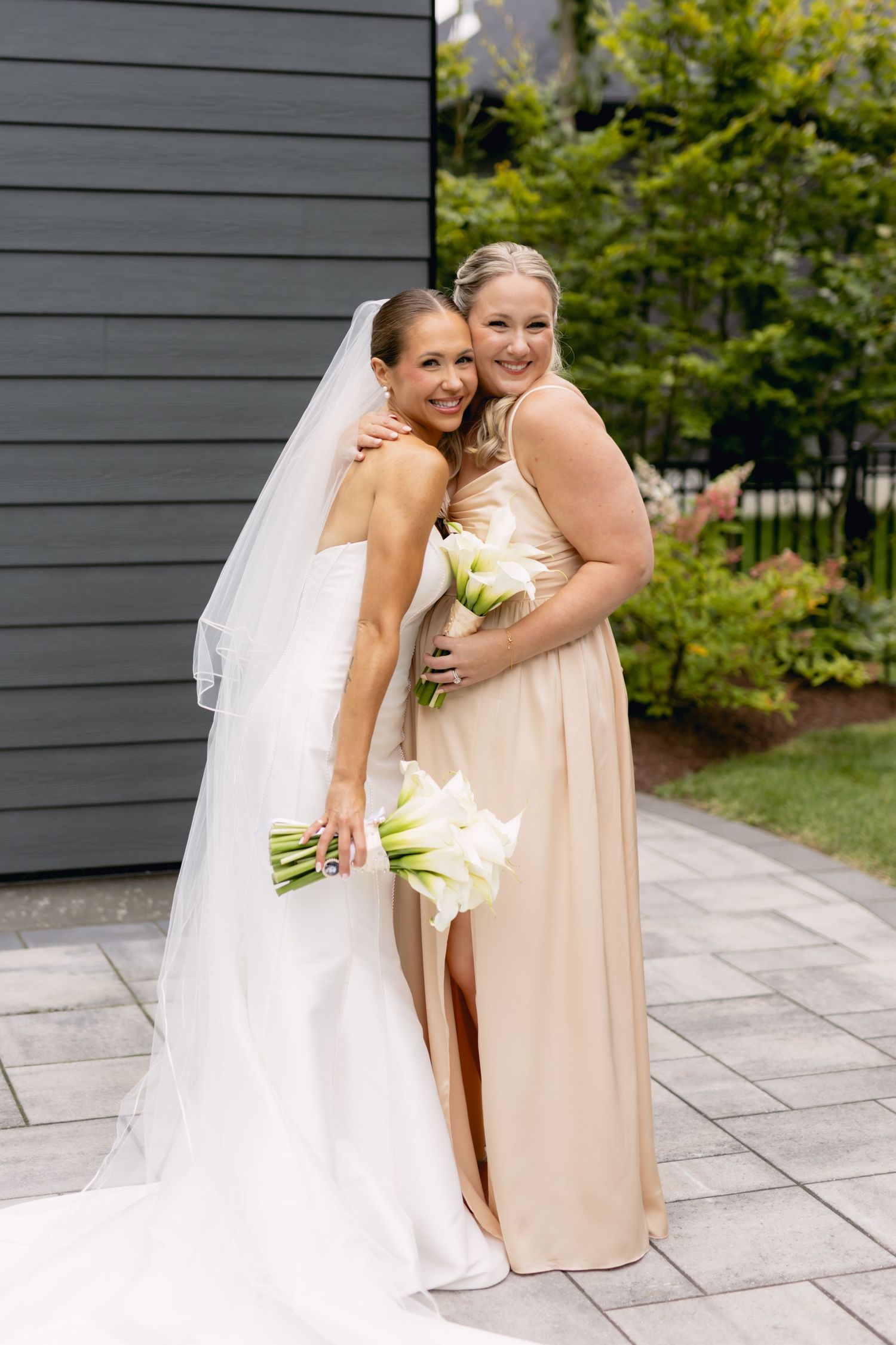 Bride in white gown and veil embraces bridesmaid in champagne dress, both holding white calla lily bouquets outdoors.