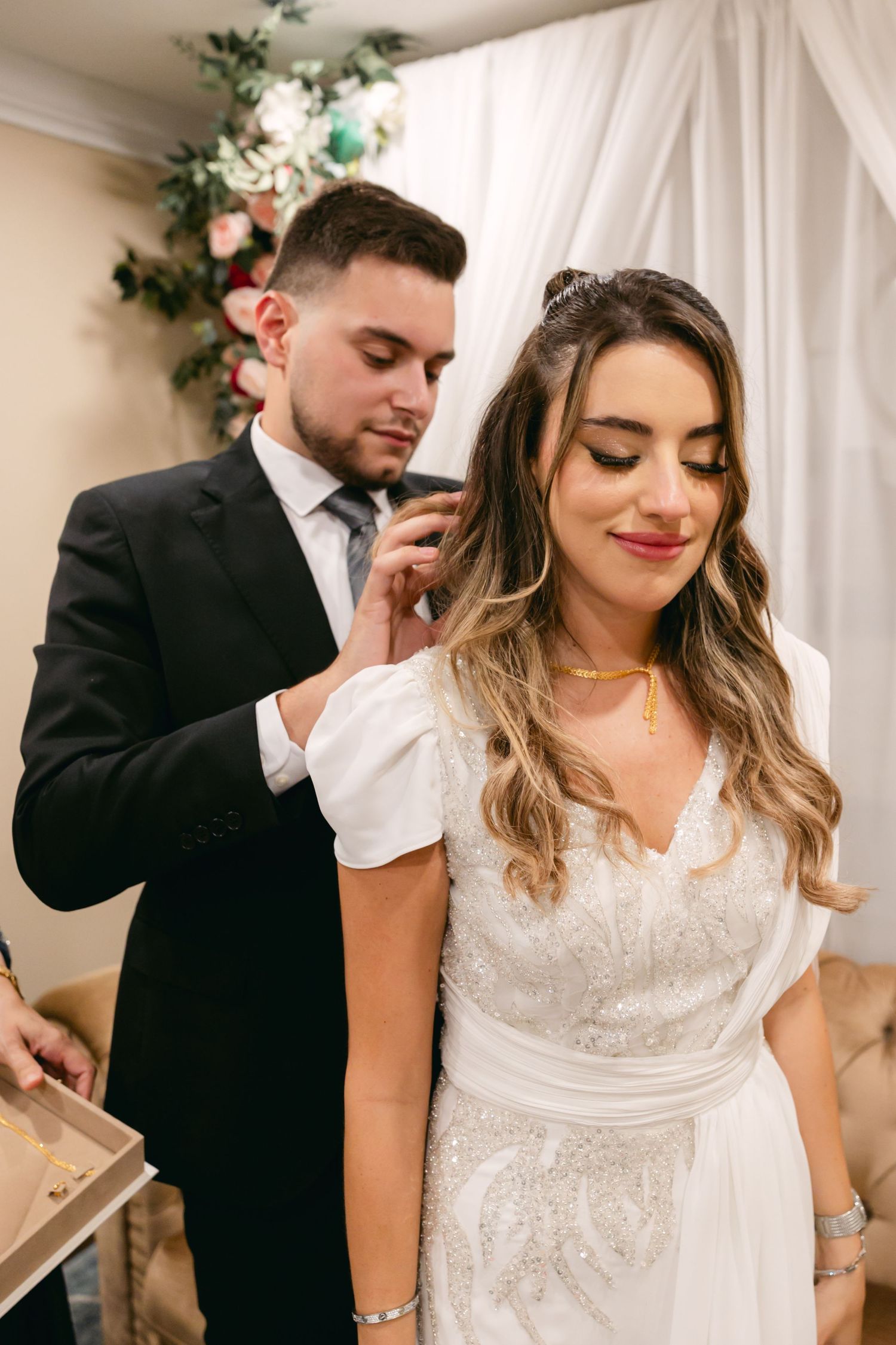 Wedding preparation scene with white drapes and floral decorations in the background.