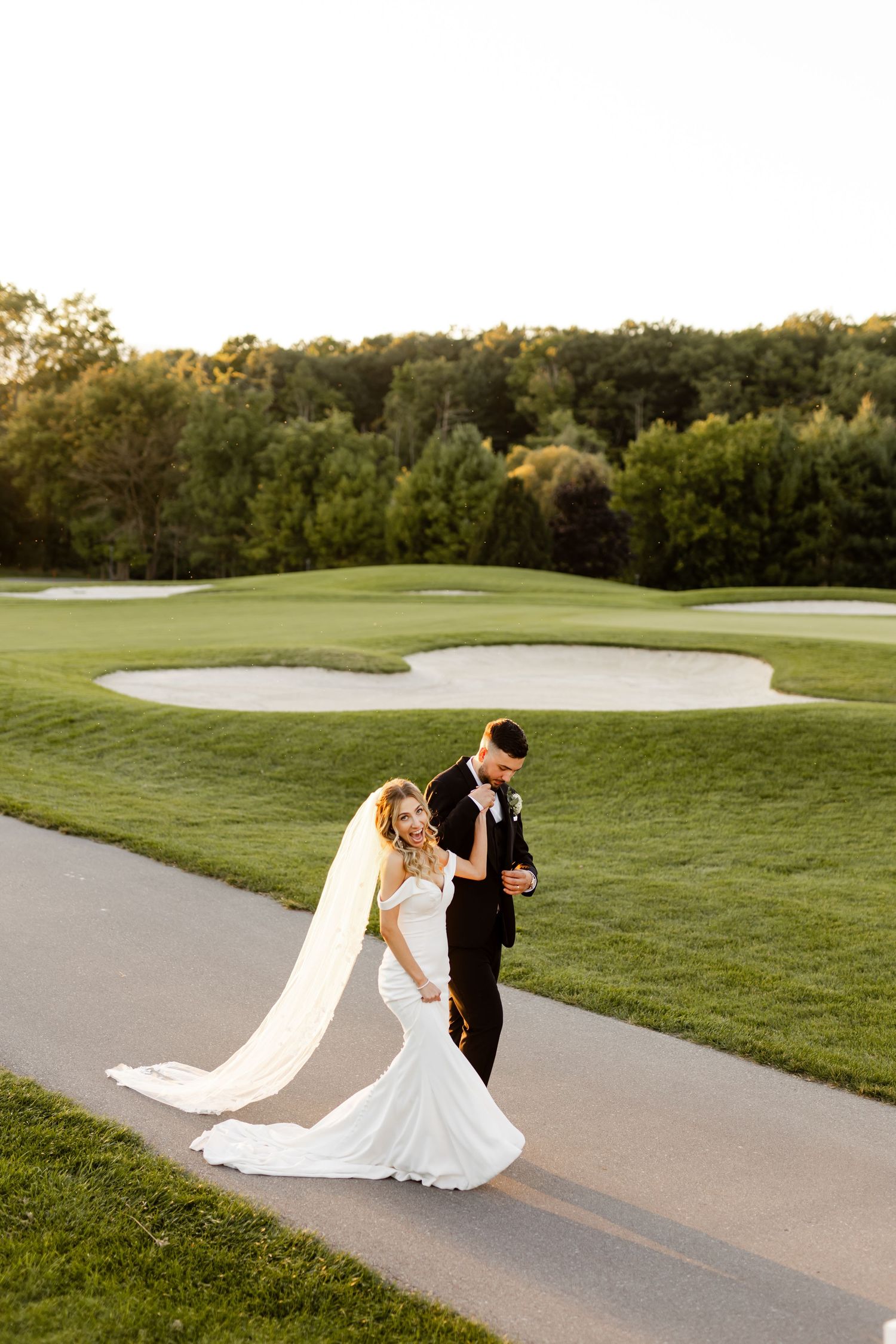 Couple in wedding attire dance together on a winding golf cart path at golden hour.
