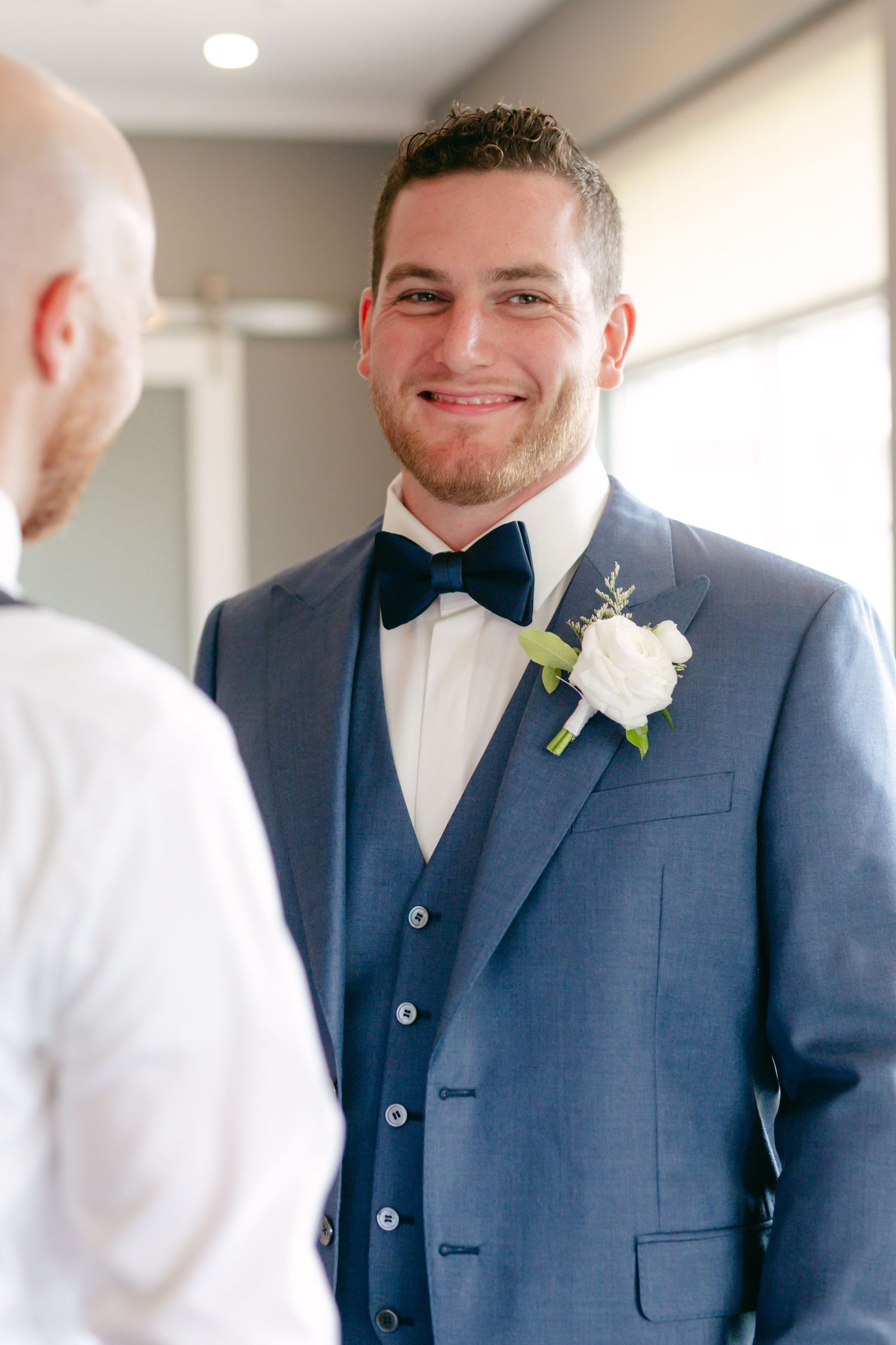 Groom in navy blue suit with white boutonniere adjusts his bow tie while getting ready for the wedding ceremony.