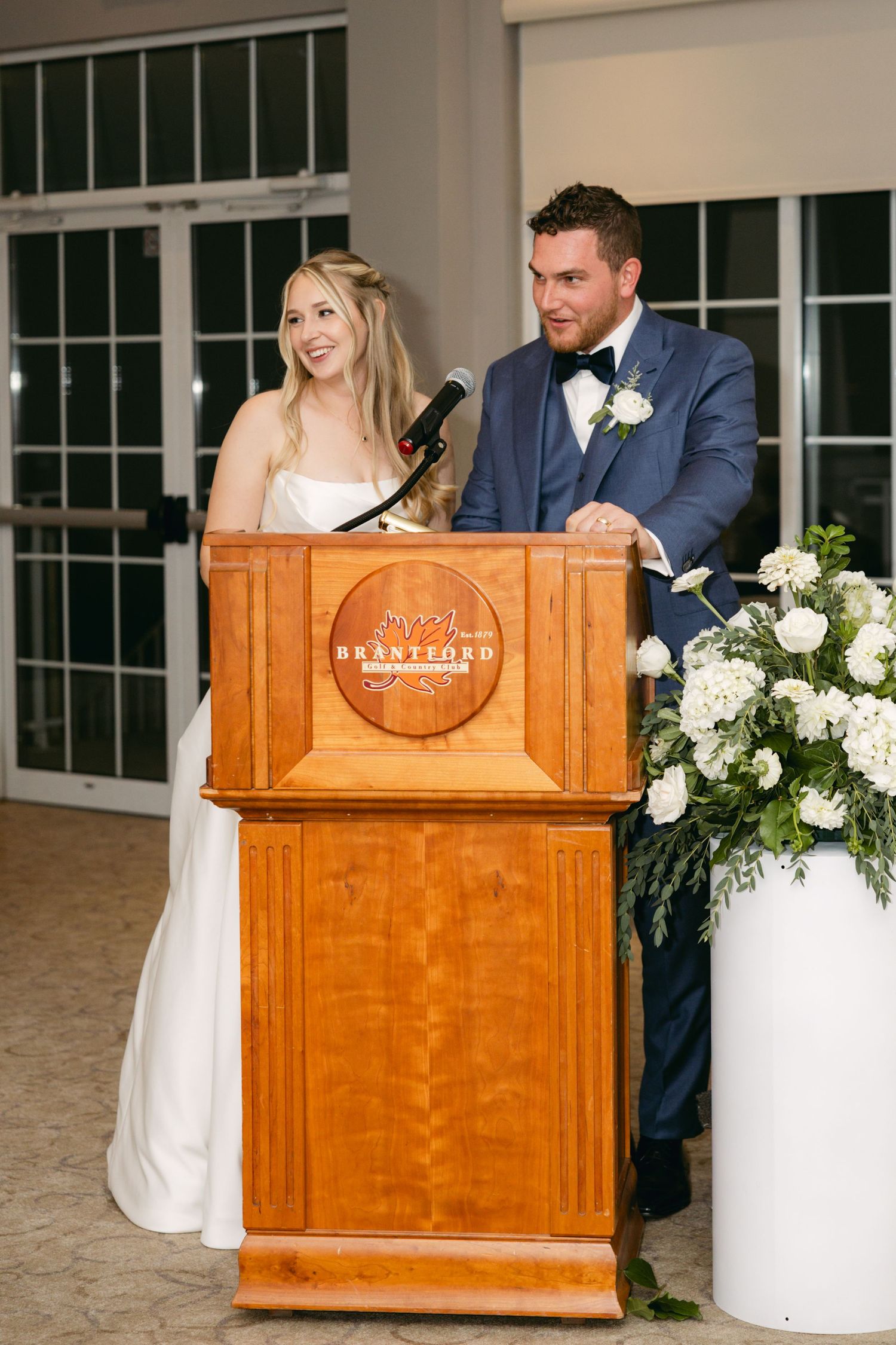 A wooden podium with white floral arrangements sits in front of a window during a wedding ceremony.