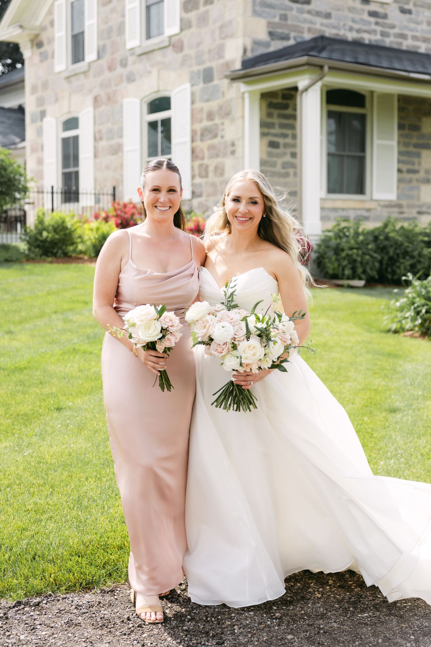 Bride in white gown and bridesmaid in pink dress pose together on green lawn.