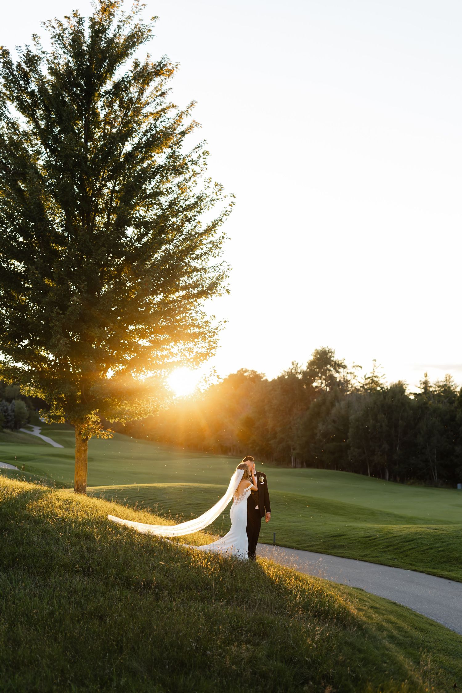 Dramatic sunset wedding portrait on a golf course with flowing veil silhouetted against golden light.