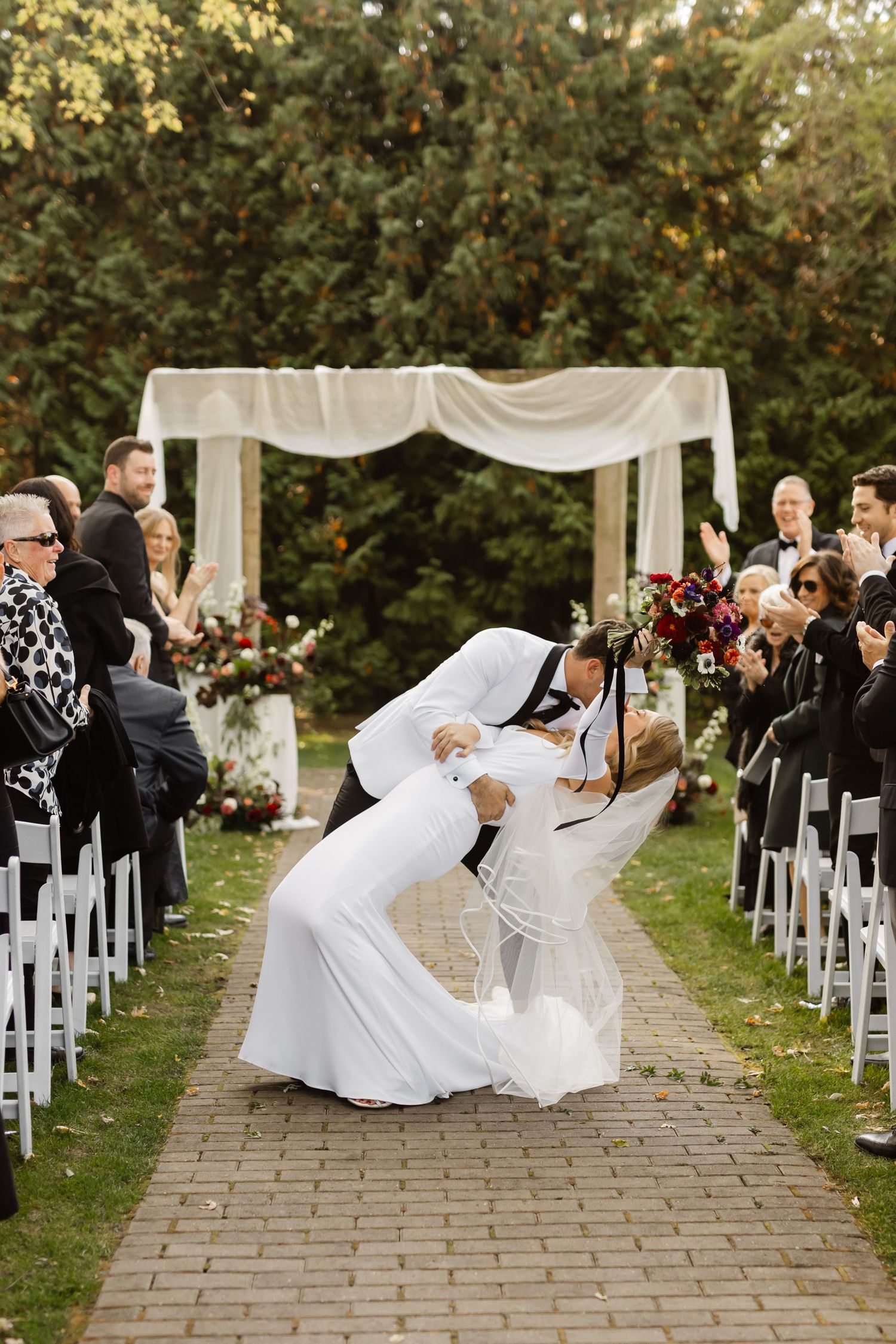 Wedding ceremony setup with white chairs and flowing veils on a stone pathway surrounded by autumn trees.