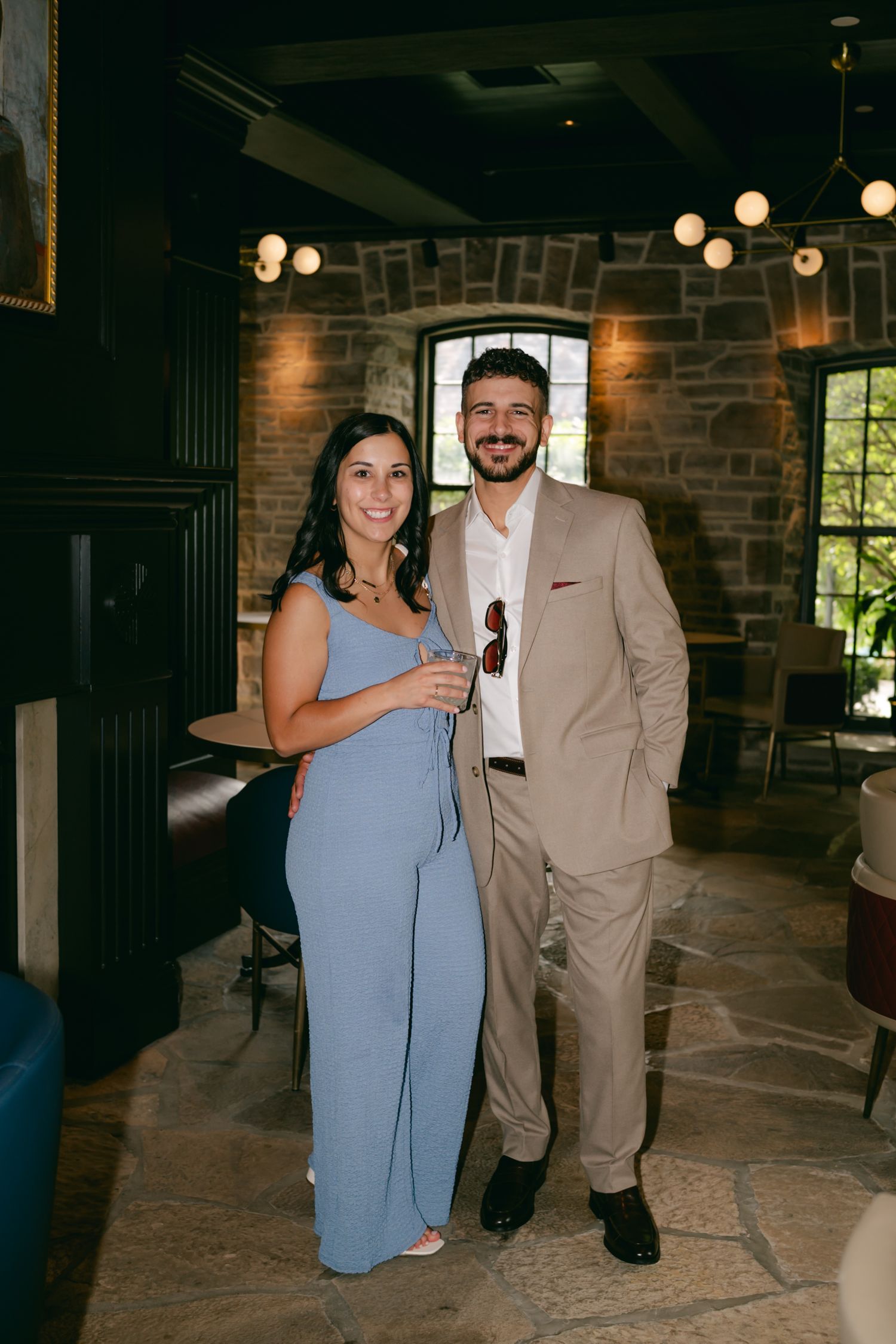Couple in blue dress and tan suit pose at rustic venue with exposed brick walls.