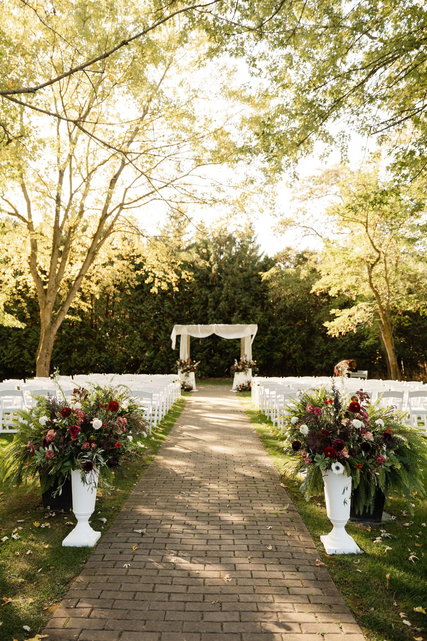 Outdoor wedding ceremony setup with white chairs and floral arrangements along brick pathway.