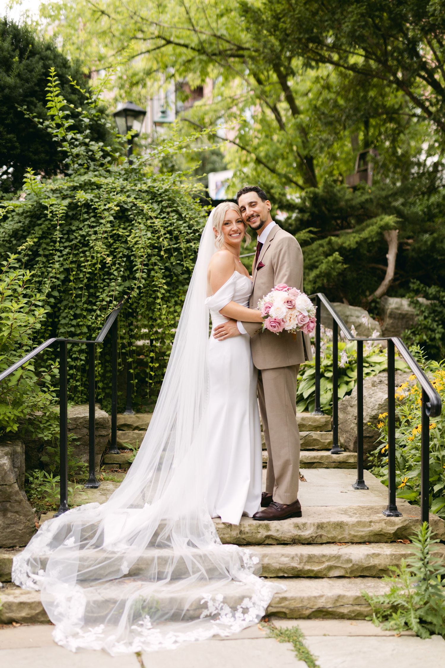 Wedding couple poses on stone steps surrounded by lush greenery in a romantic outdoor setting.