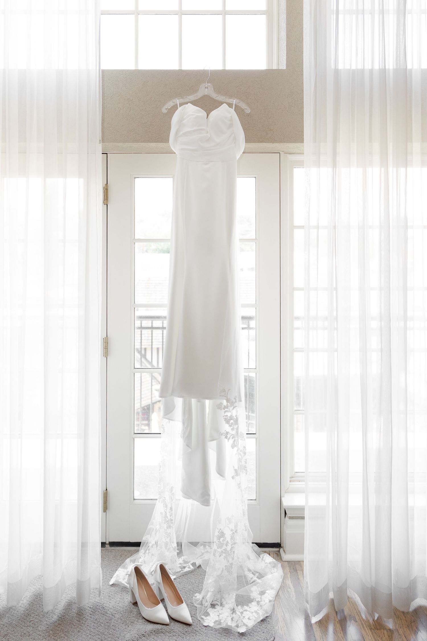White wedding dress and shoes hanging by window with soft natural light and sheer curtains.