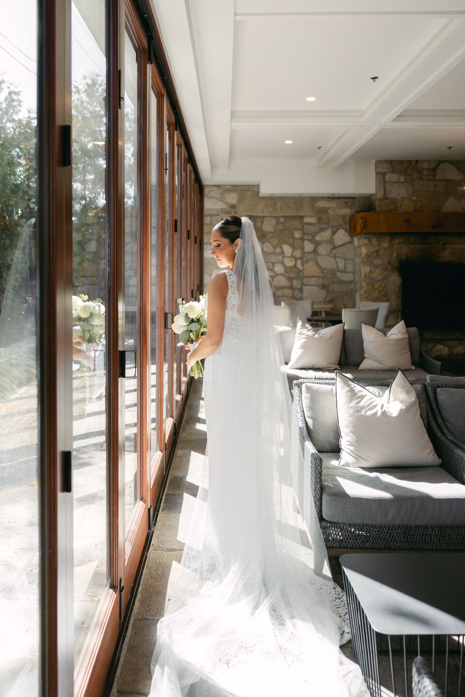 A bride in a flowing white gown stands in a bright room with floor-to-ceiling windows.