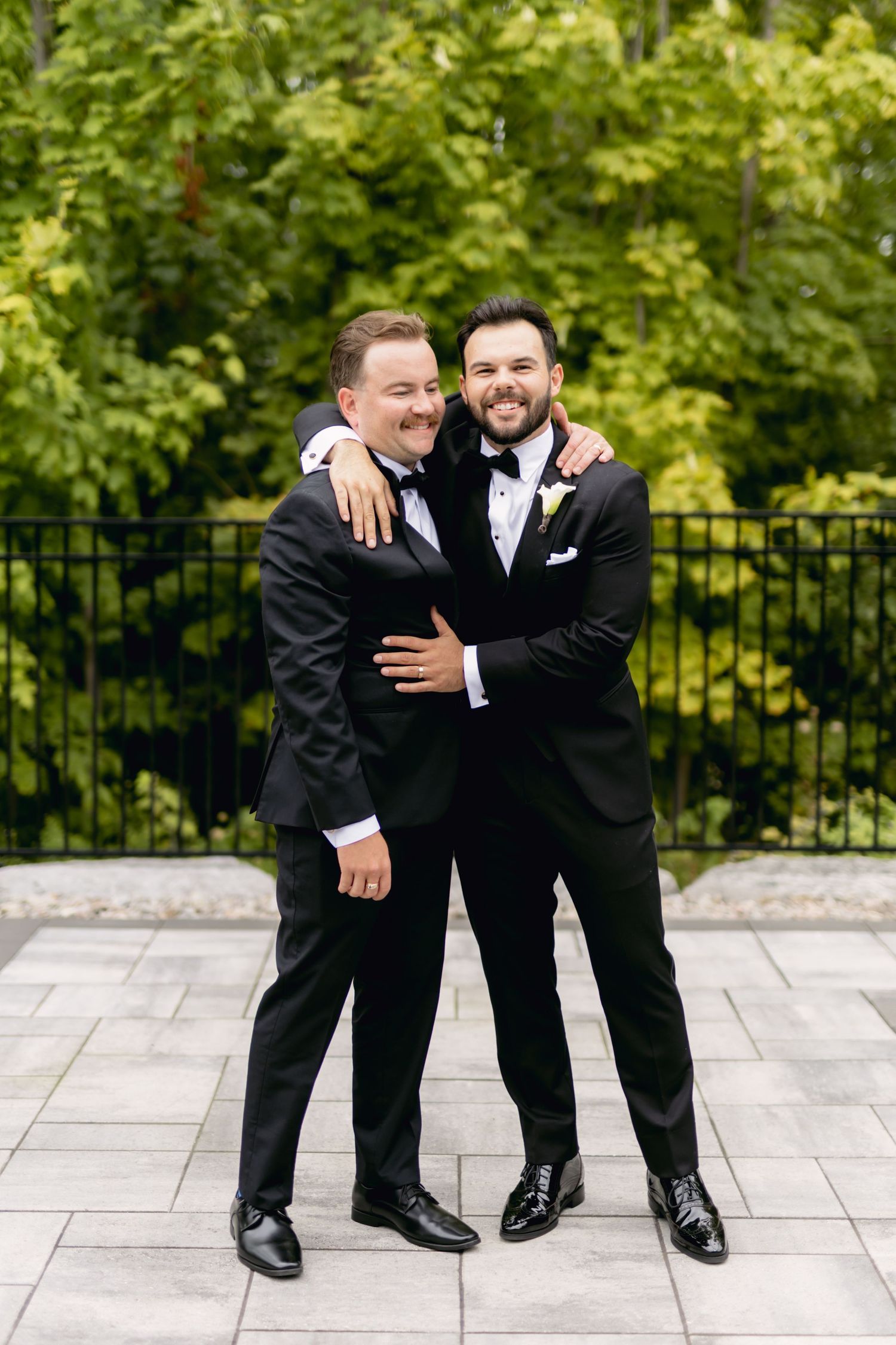 Two groomsmen in matching black tuxedos share a warm embrace outdoors with lush green trees in the background.