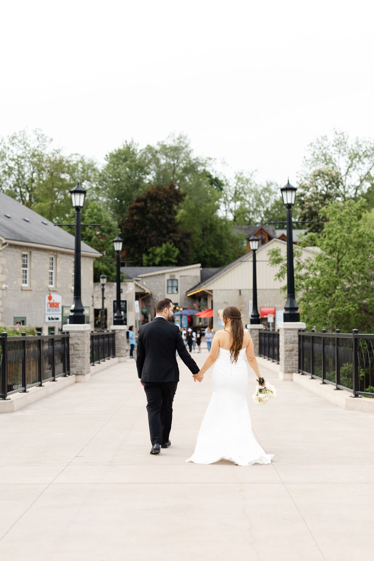 Bride and groom walk hand in hand across a bridge surrounded by lamp posts and lush green trees.
