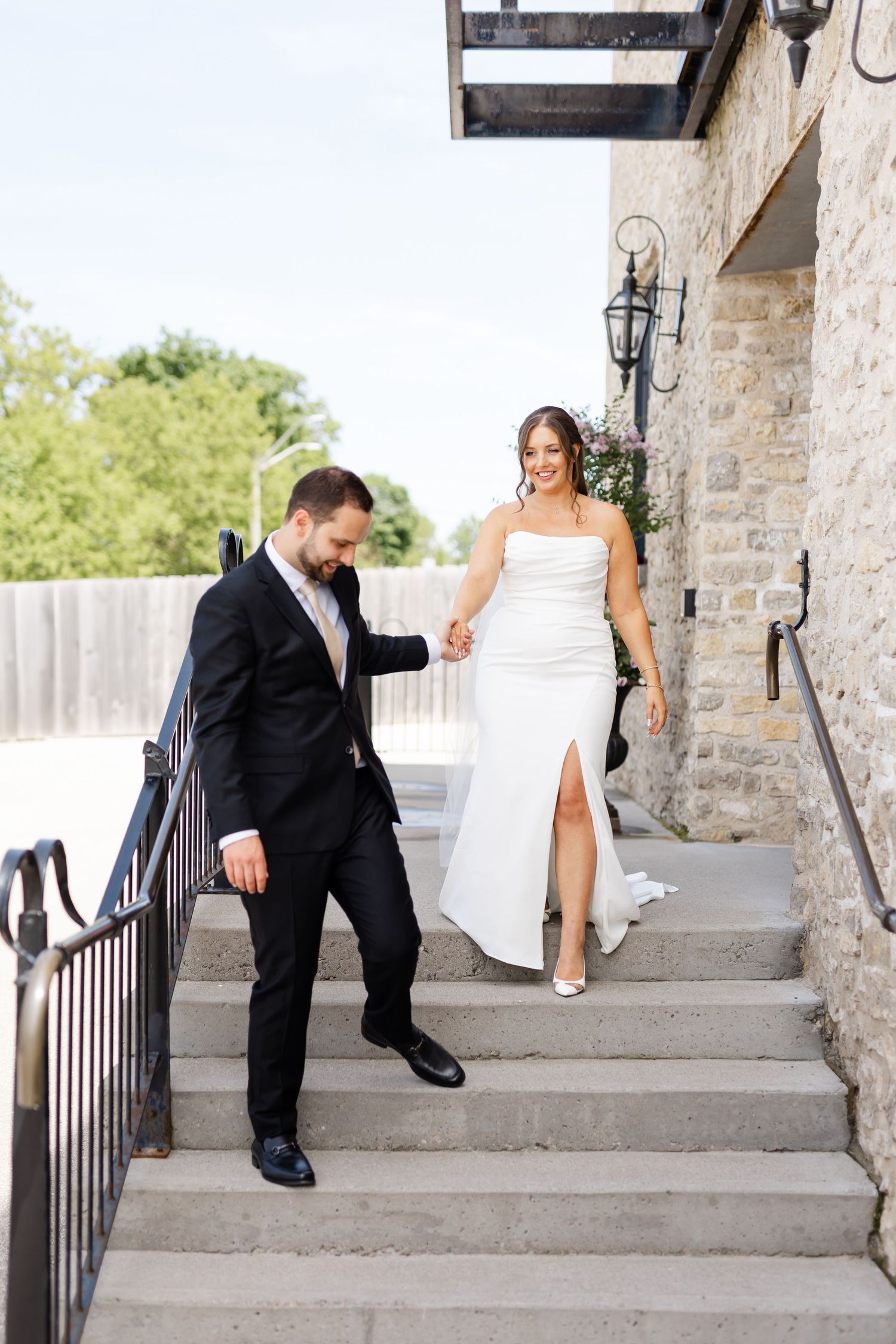 Groom helps bride descend stone steps at outdoor wedding venue, bride wearing strapless white gown with slit.