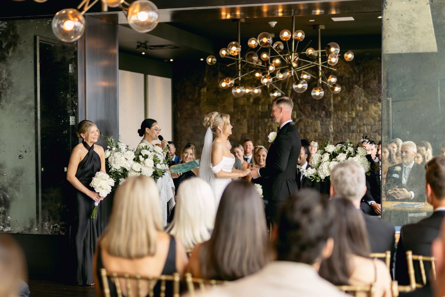Elegant indoor wedding ceremony with bride and groom at altar, gold chandelier, white floral arrangements, and seated guests.