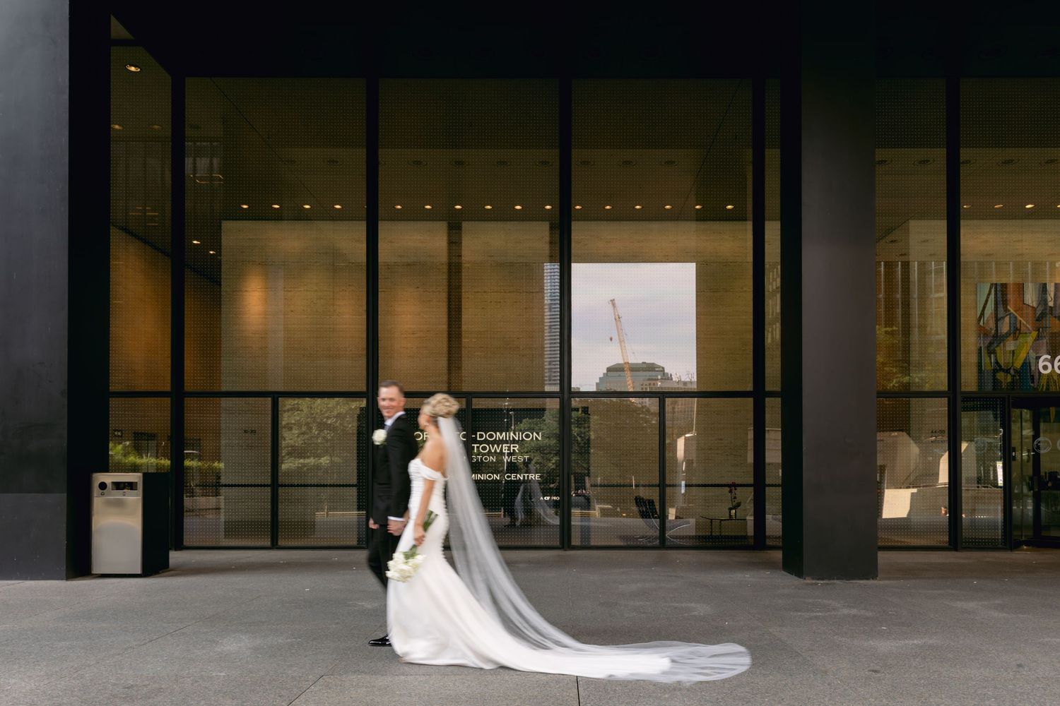 Bride in flowing white gown with cathedral veil walks with groom outside modern glass building.