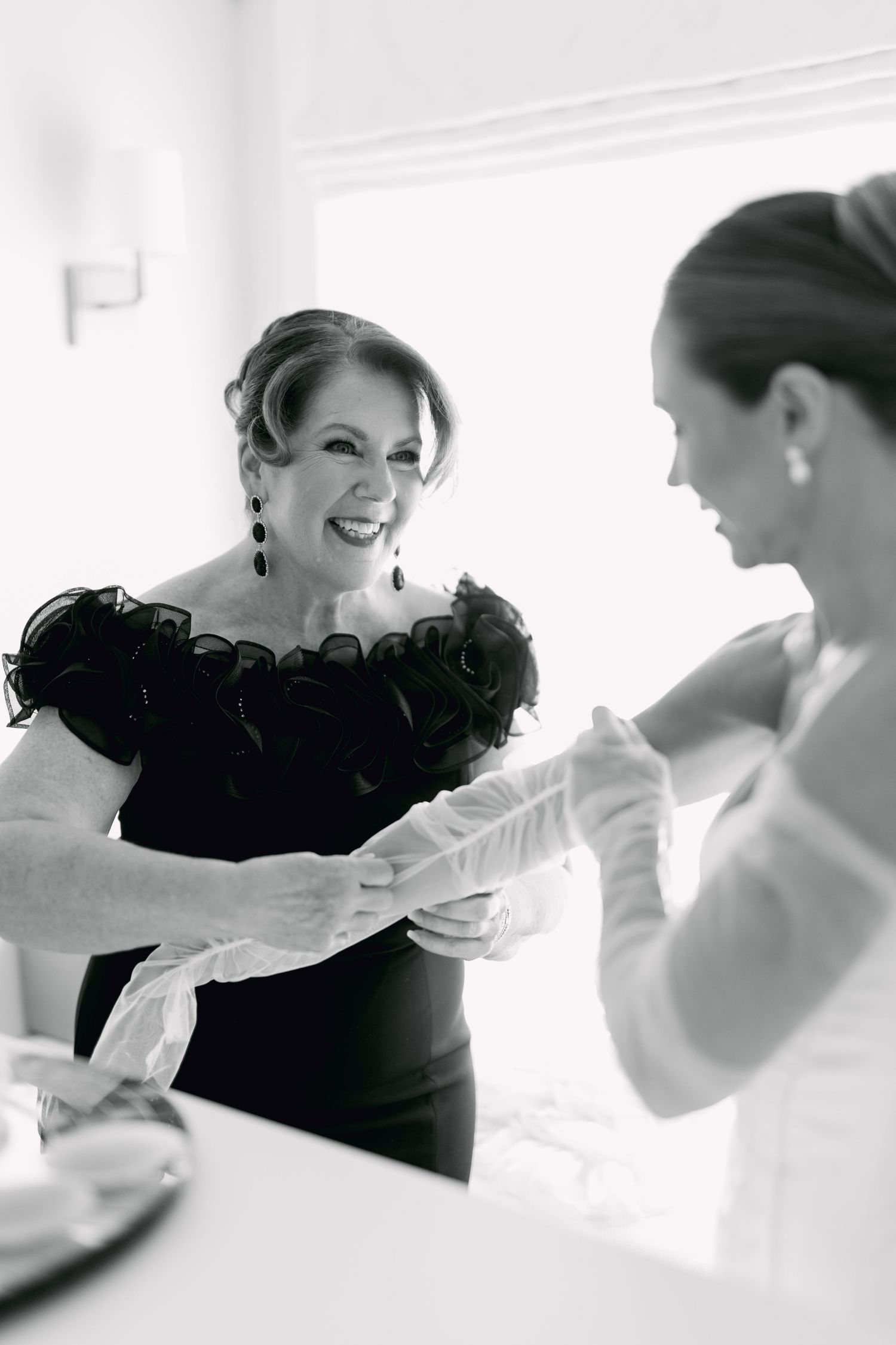 Mother helping bride with gloves during wedding preparations in black and white photo.