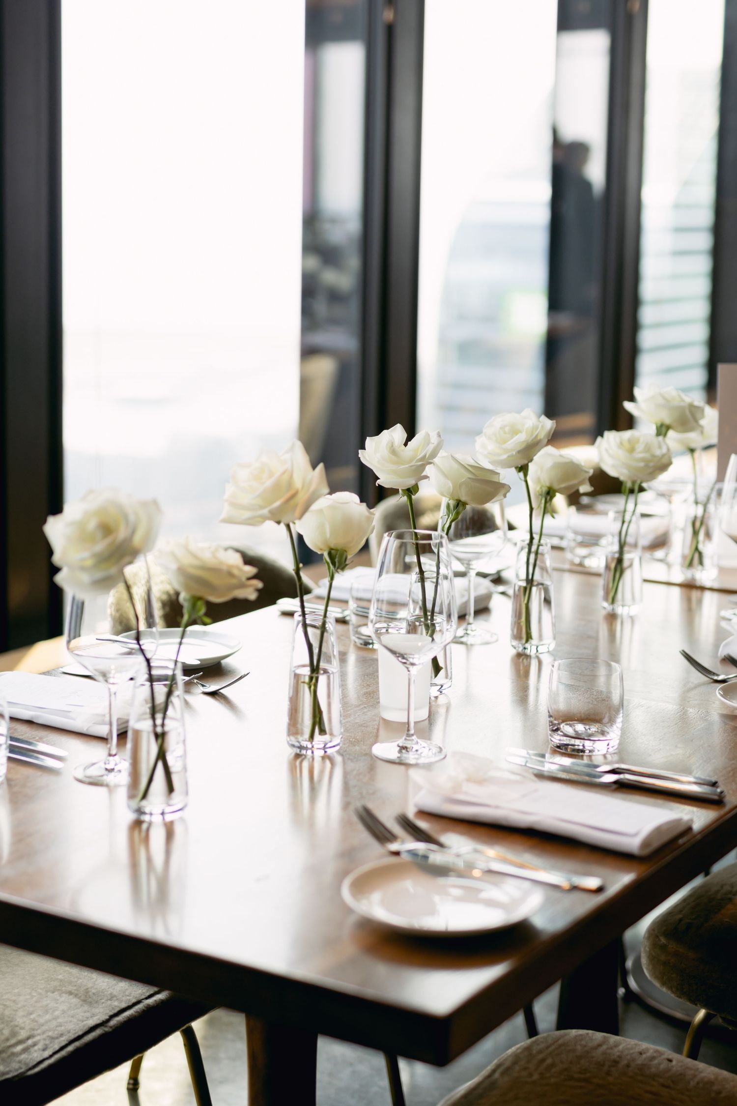 Elegant wedding reception table with white roses in glass vases, crystal glassware, and linen napkins by large windows.