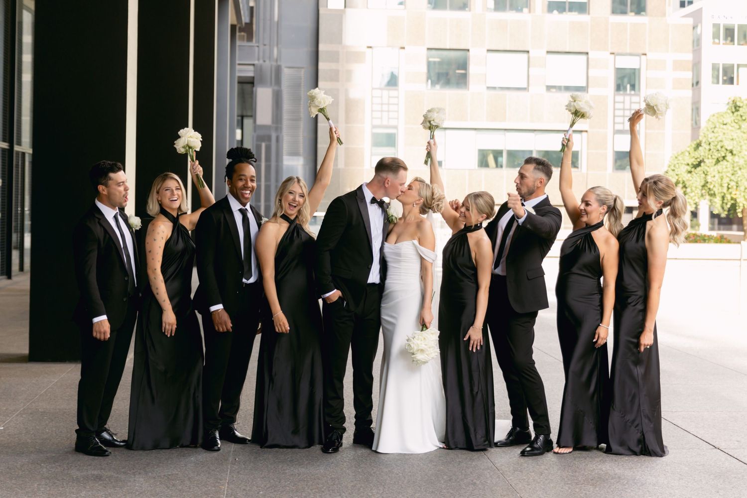 Wedding party in black attire celebrates outdoors as bride and groom kiss, bridesmaids raising bouquets in cheer.