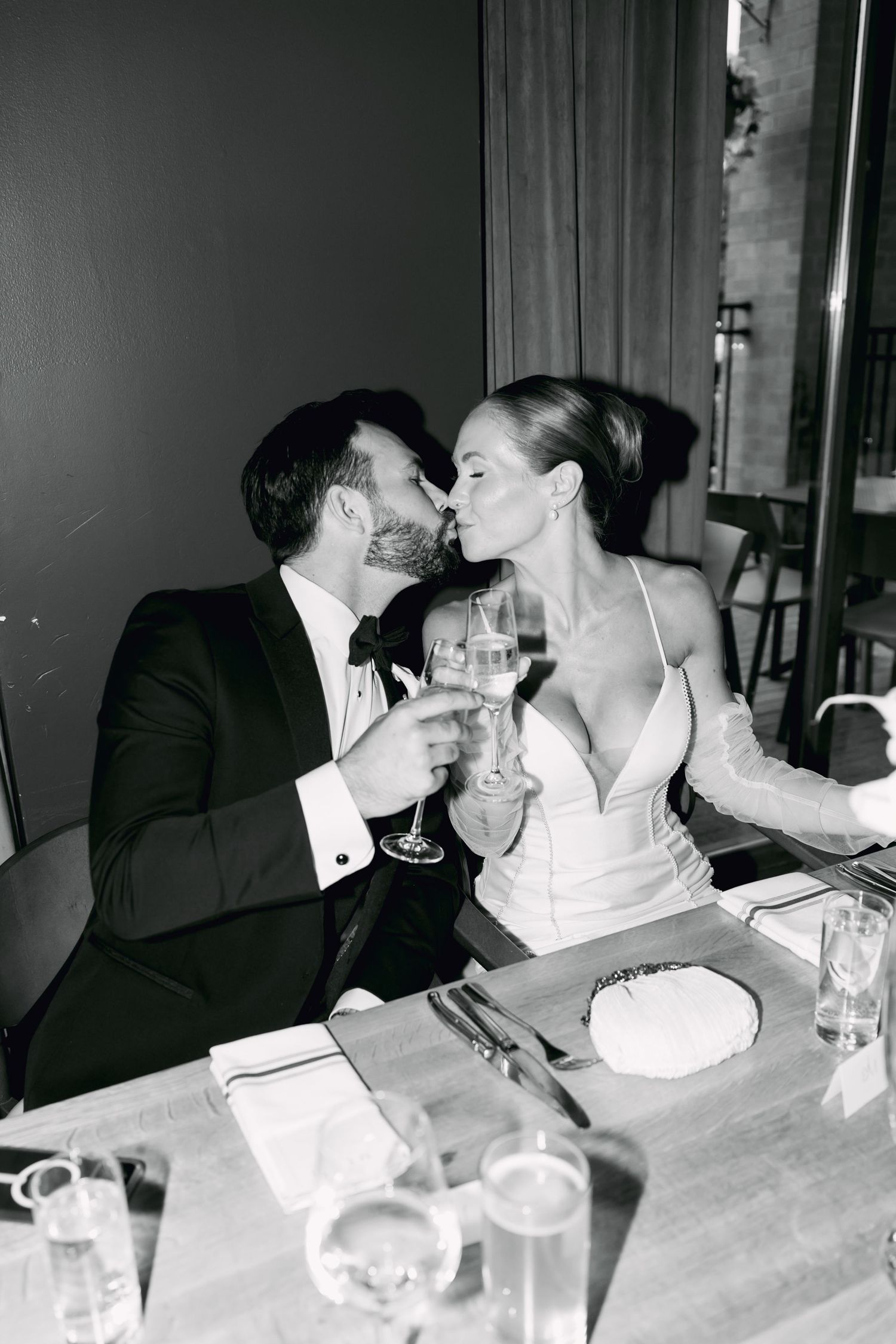 Black and white photo of bride and groom kissing while clinking champagne glasses at wedding reception table.