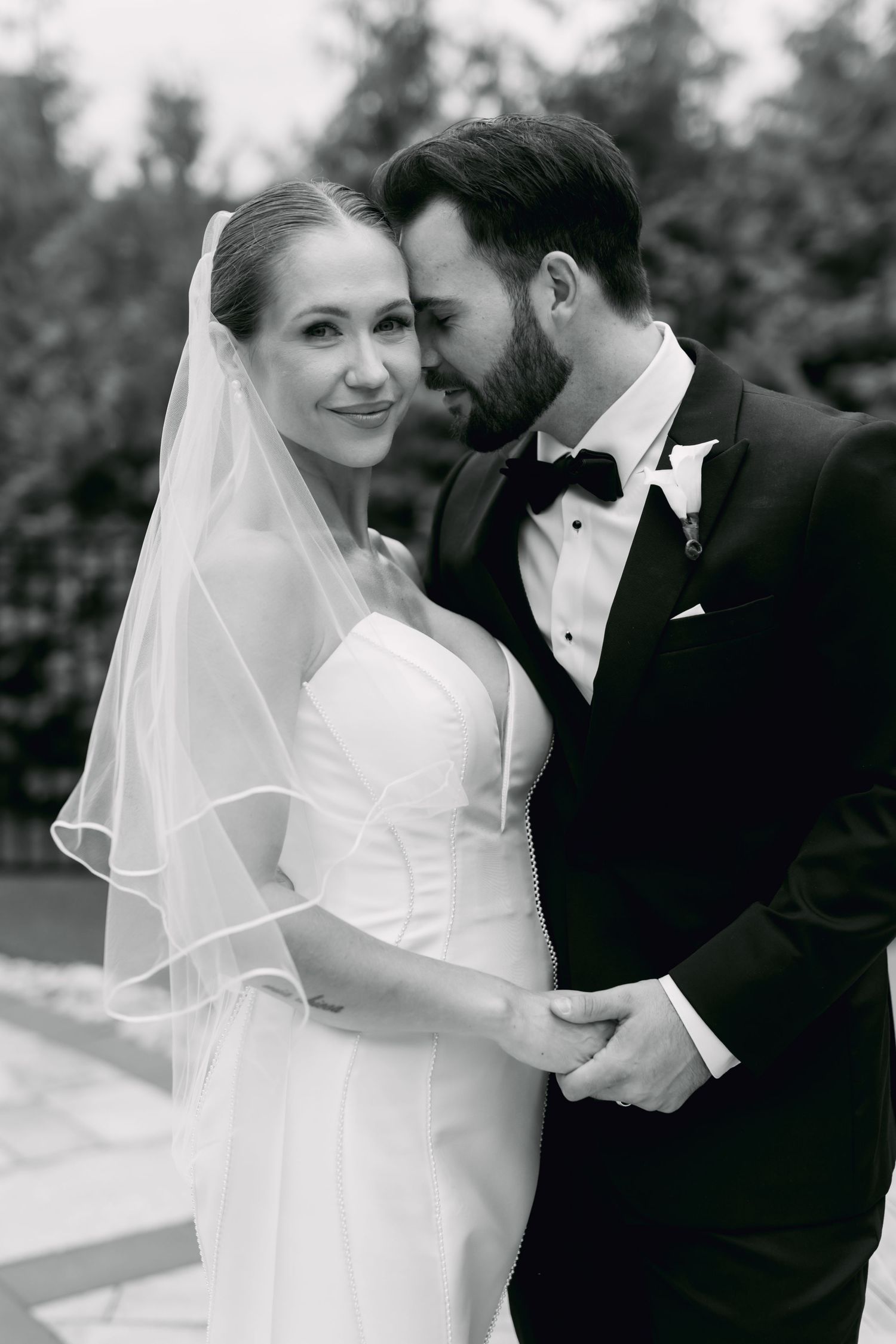 Black and white romantic portrait of bride in veil and groom in tuxedo embracing outdoors.