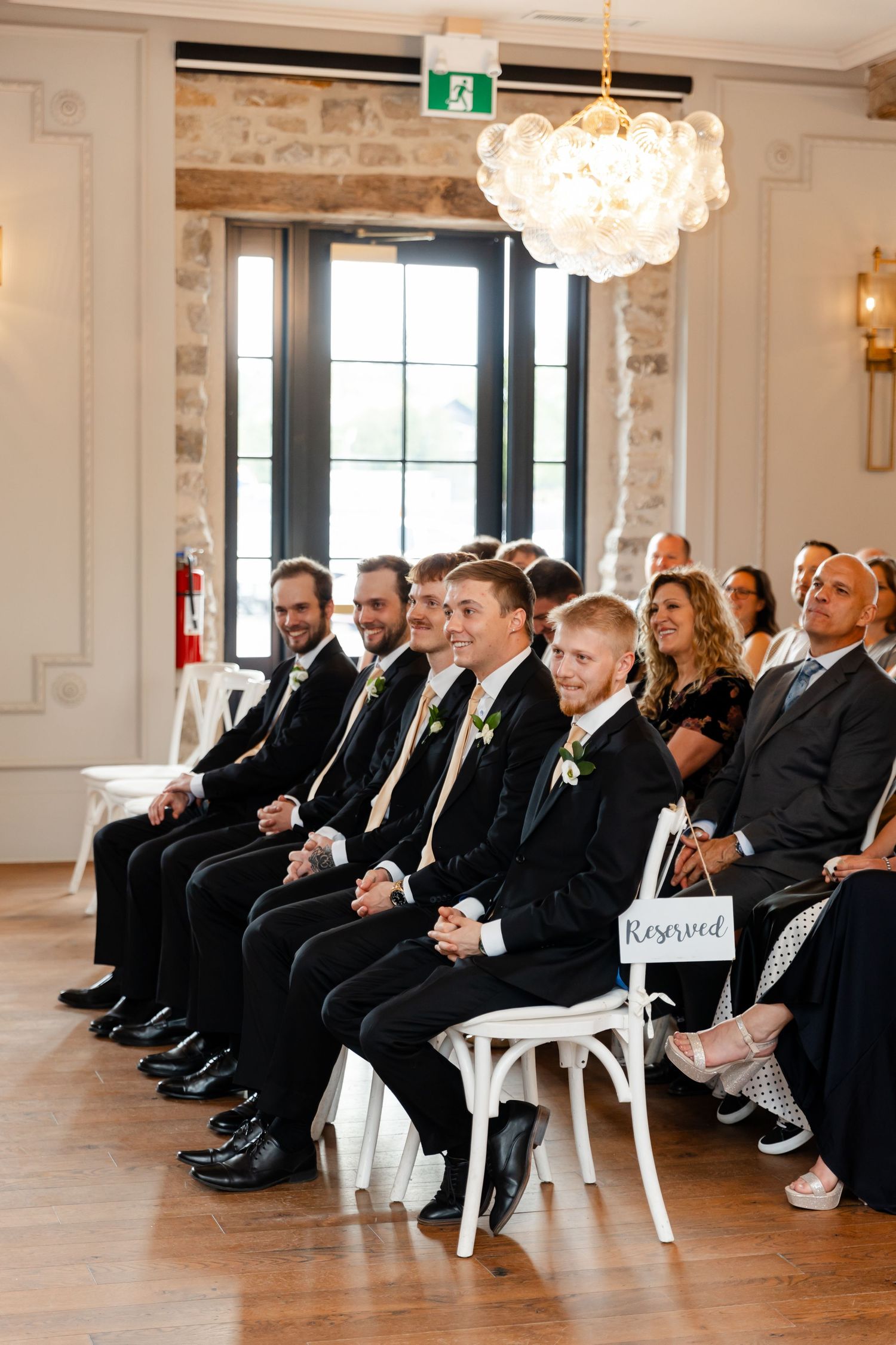 Groomsmen in black suits with boutonnieres sit smiling in reserved seats during indoor wedding ceremony.