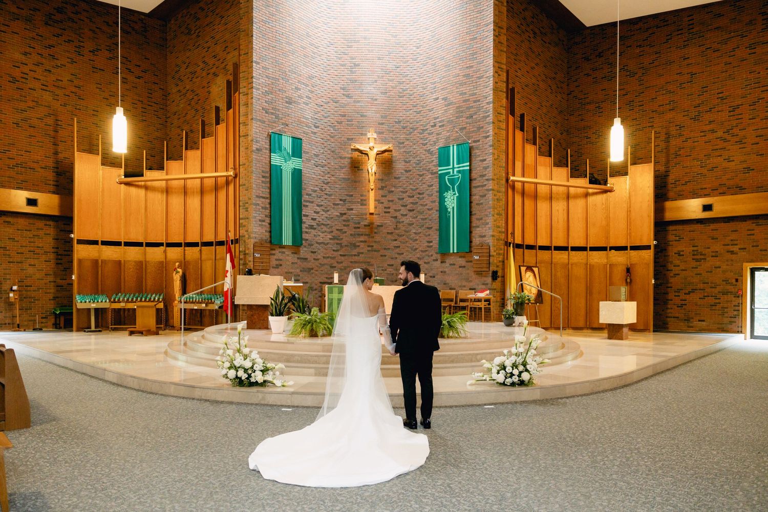 Bride and groom stand at the altar of a modern Catholic church with green banners and a golden crucifix.