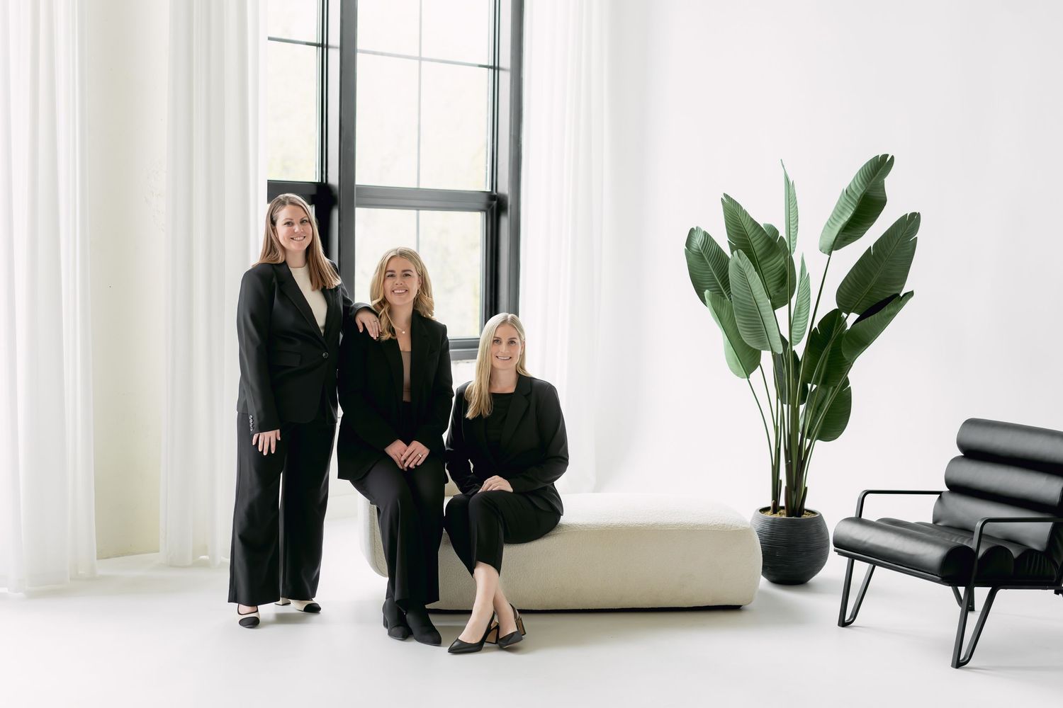Three women in black outfits pose in a bright modern studio with large windows and a tropical plant.