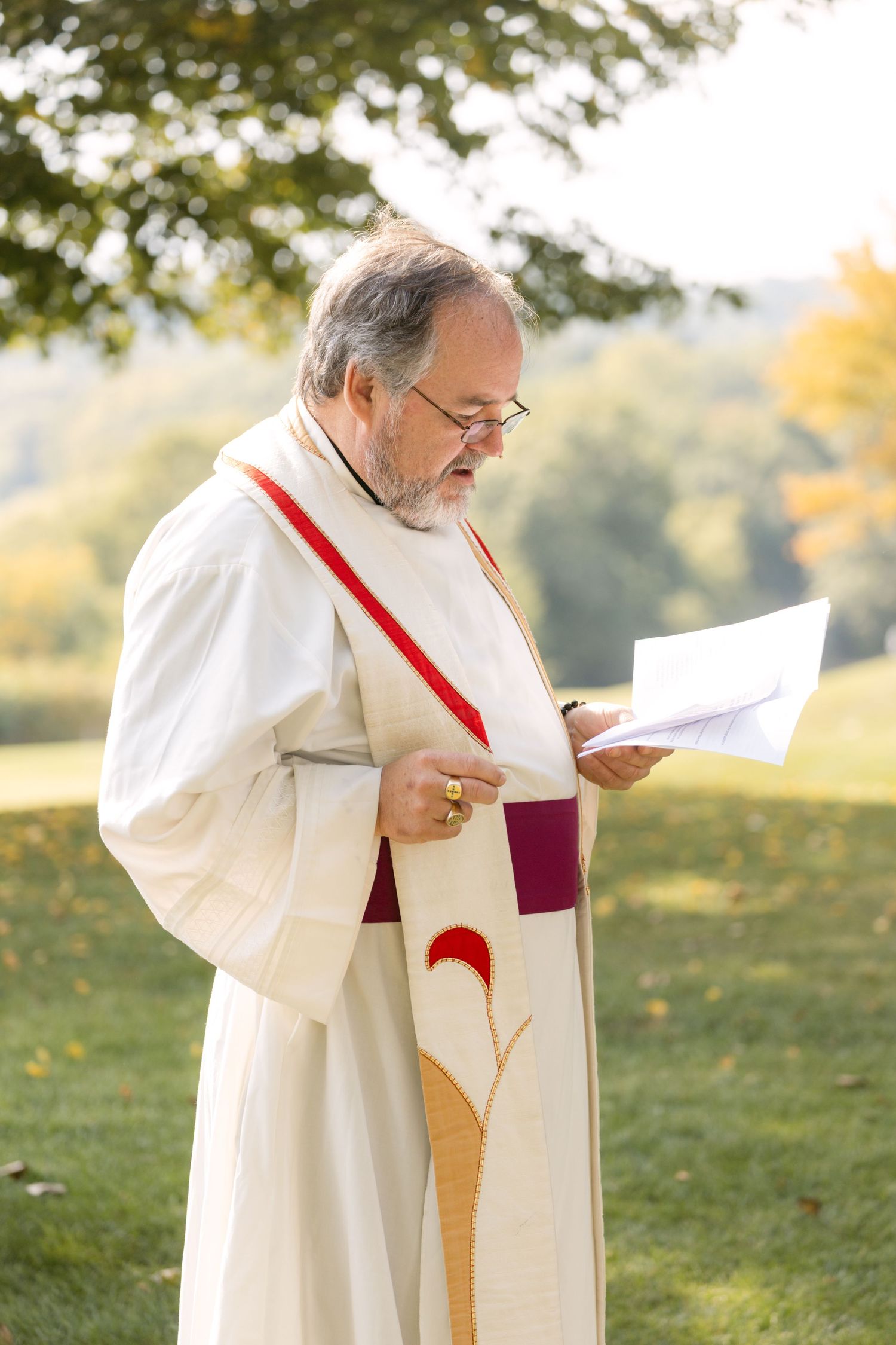 Religious officiant in white robes with red stole reading from ceremony text outdoors.