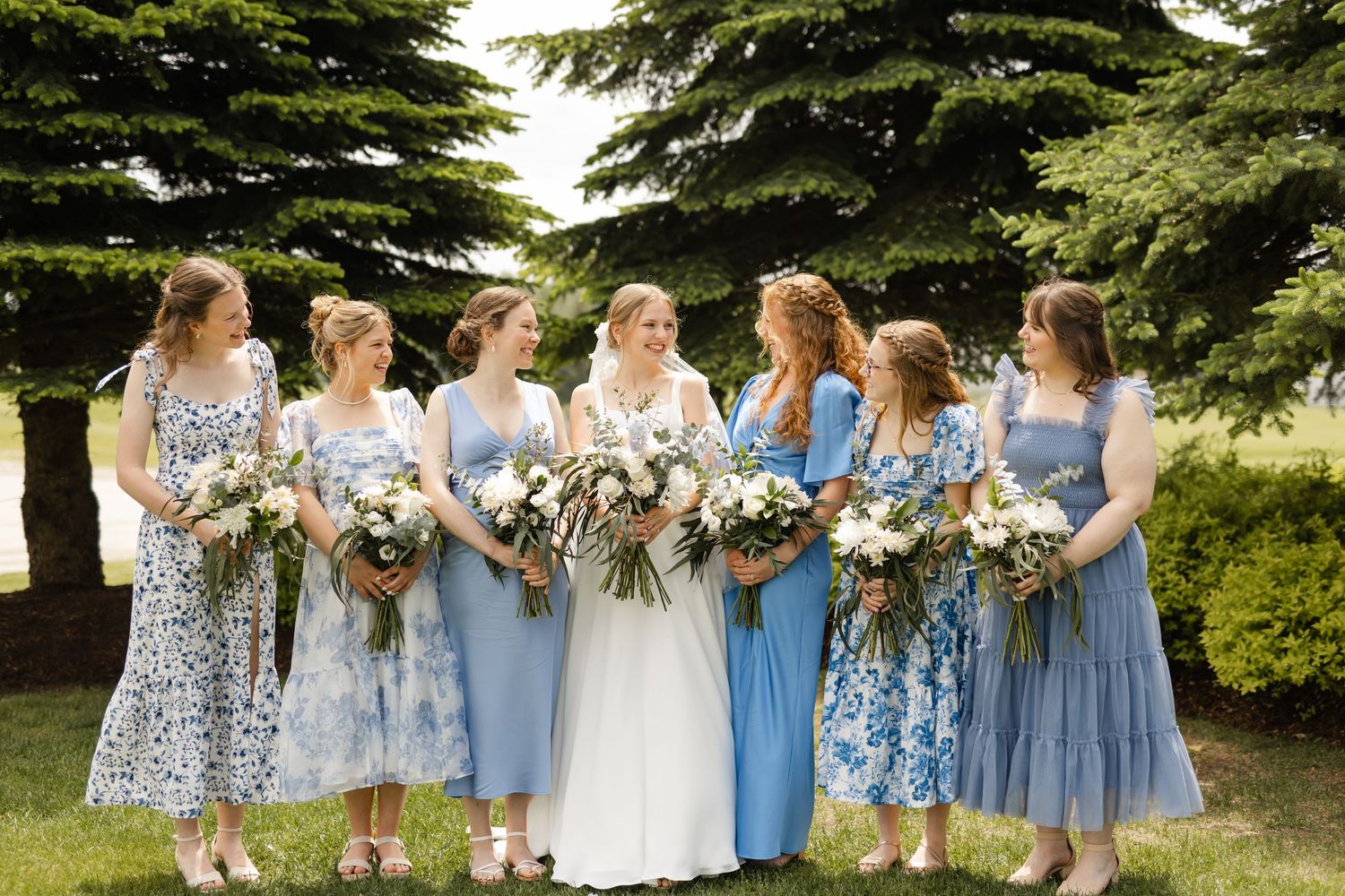 Wedding party in blue and floral dresses standing together outdoors with bouquets.