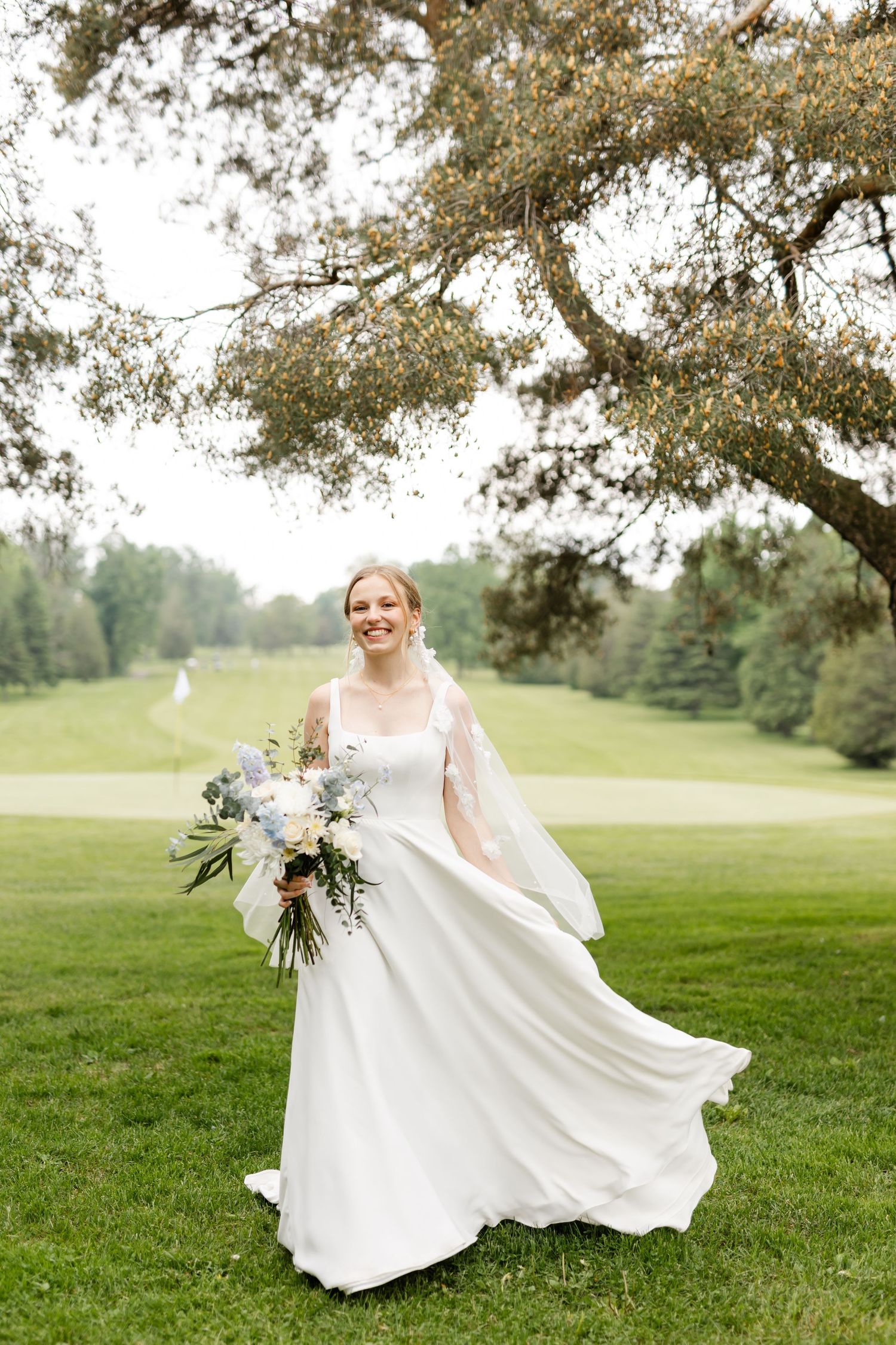 Bride's dress flows gracefully as she stands on golf course holding floral bouquet.