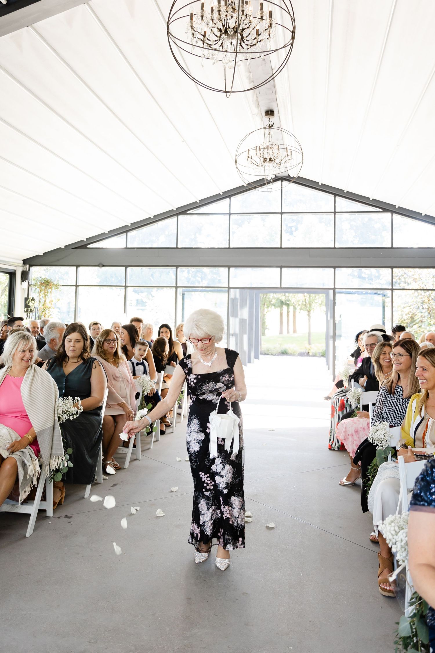 Wedding ceremony setup in white tent with crystal chandeliers and rows of white chairs.