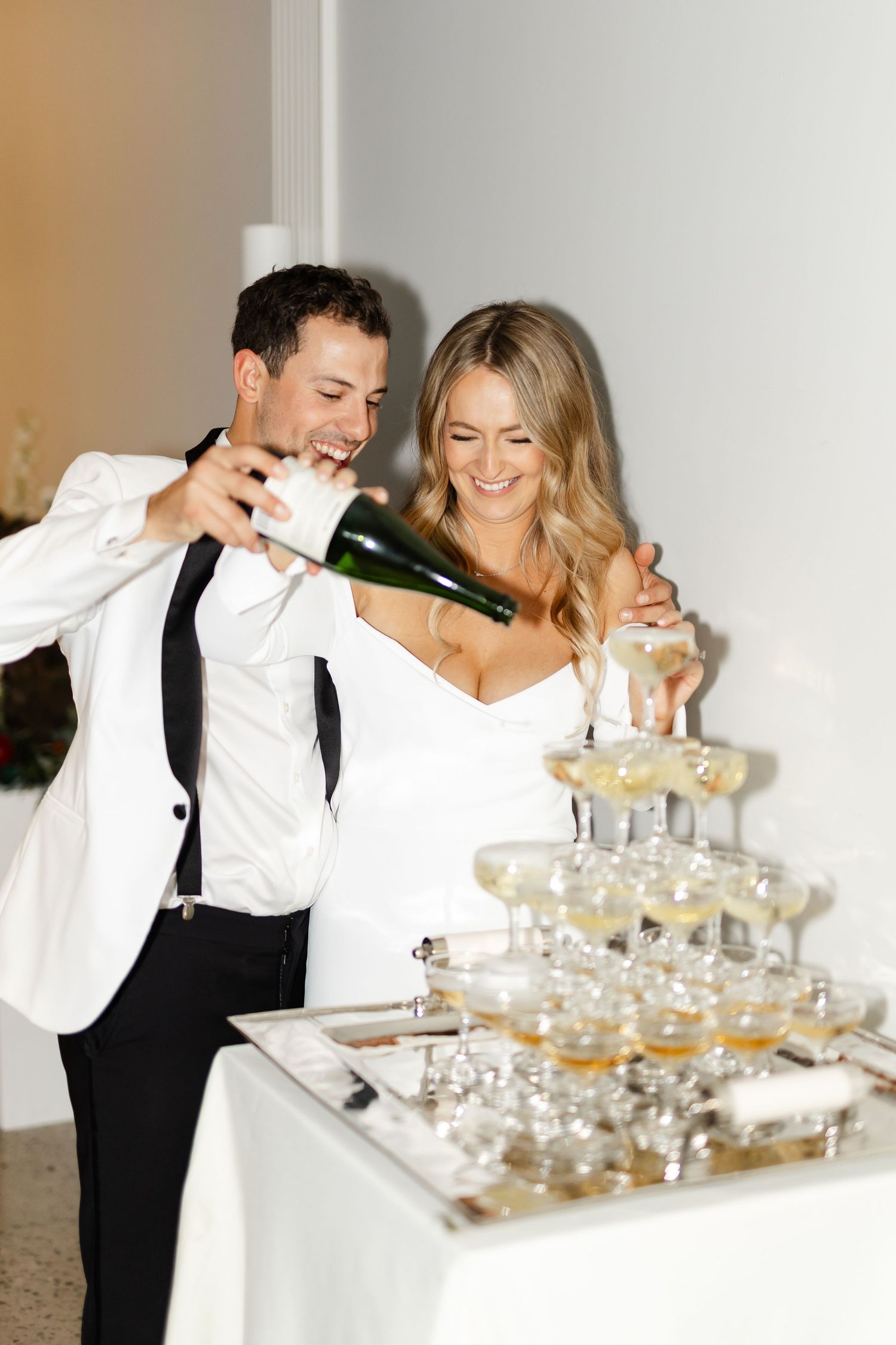 Couple sharing a joyful moment while pouring champagne into stacked glasses at their reception.