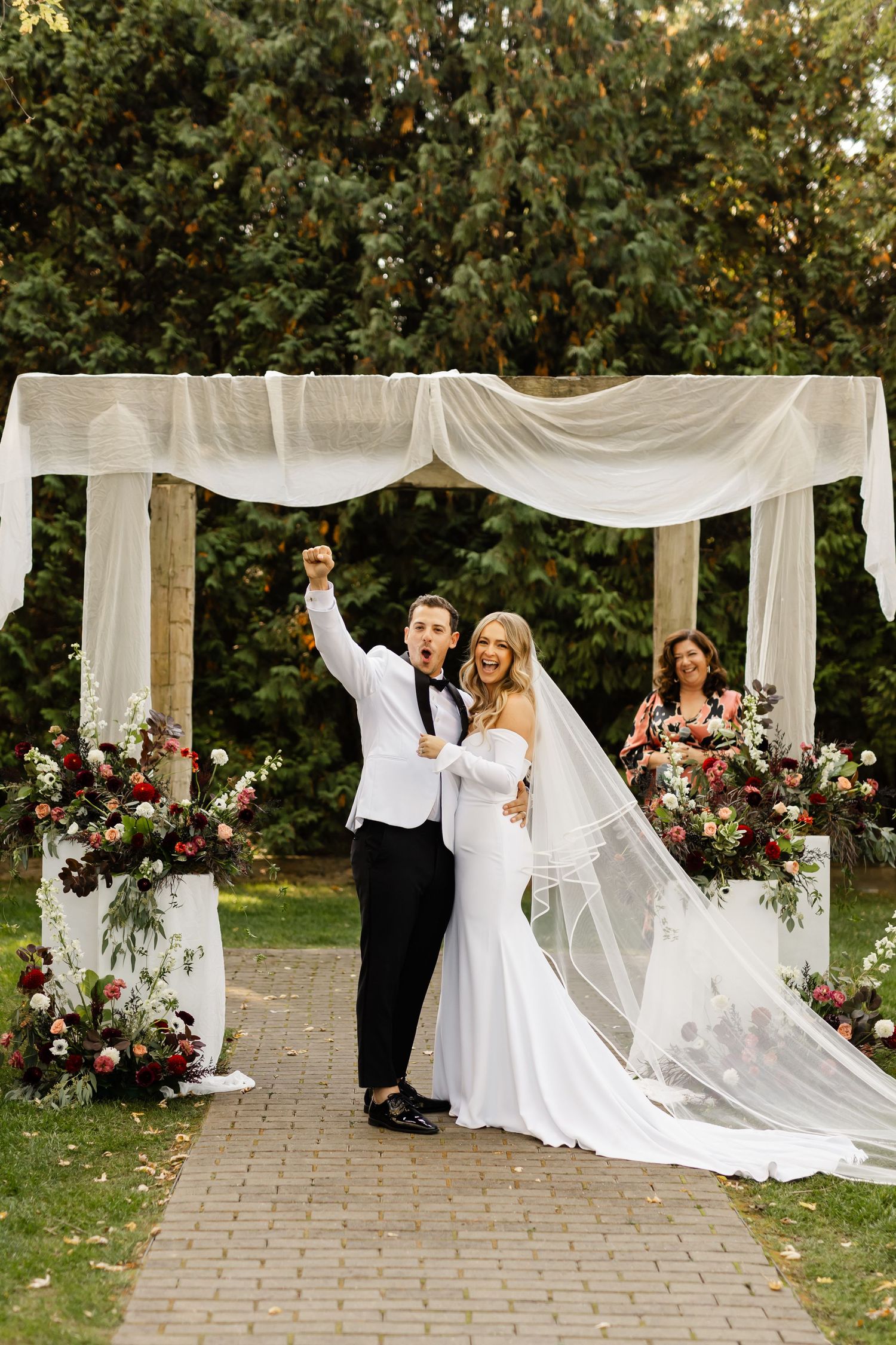 Wedding ceremony setup with white draped arch and floral arrangements on a stone pathway surrounded by autumn trees.